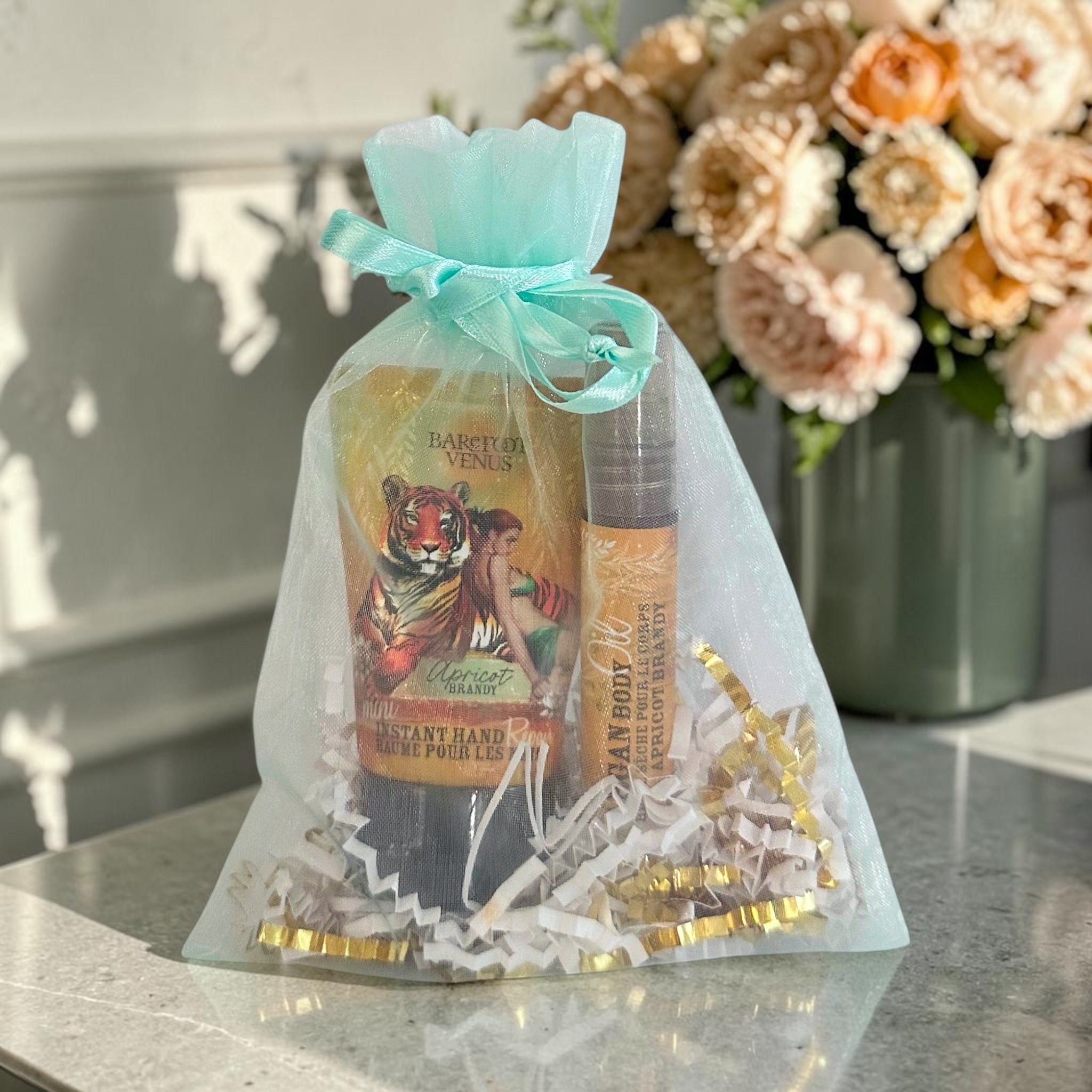 A gift set in an organza bag tied with a mint ribbon, containing a Barefoot Venus hand cream tube and a body spray bottle, filled with white and gold crinkle paper on a marble surface with flowers in the background.