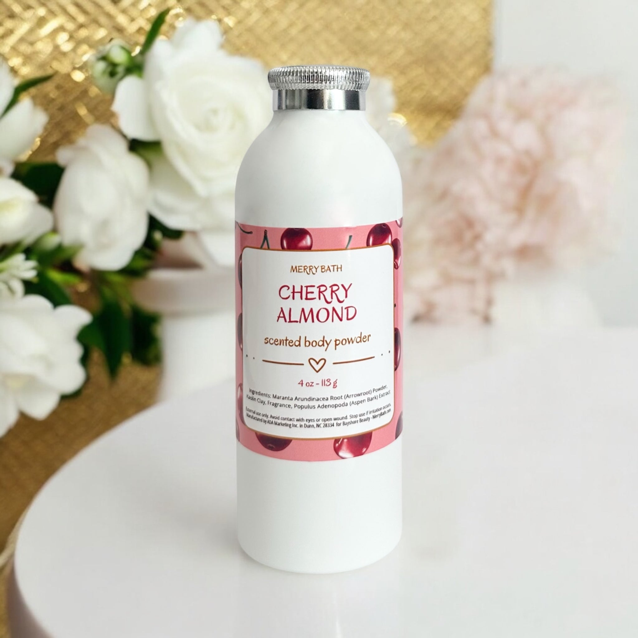 White bottle of cherry almond scented body powder with a pink label on a white surface with flowers in the background.