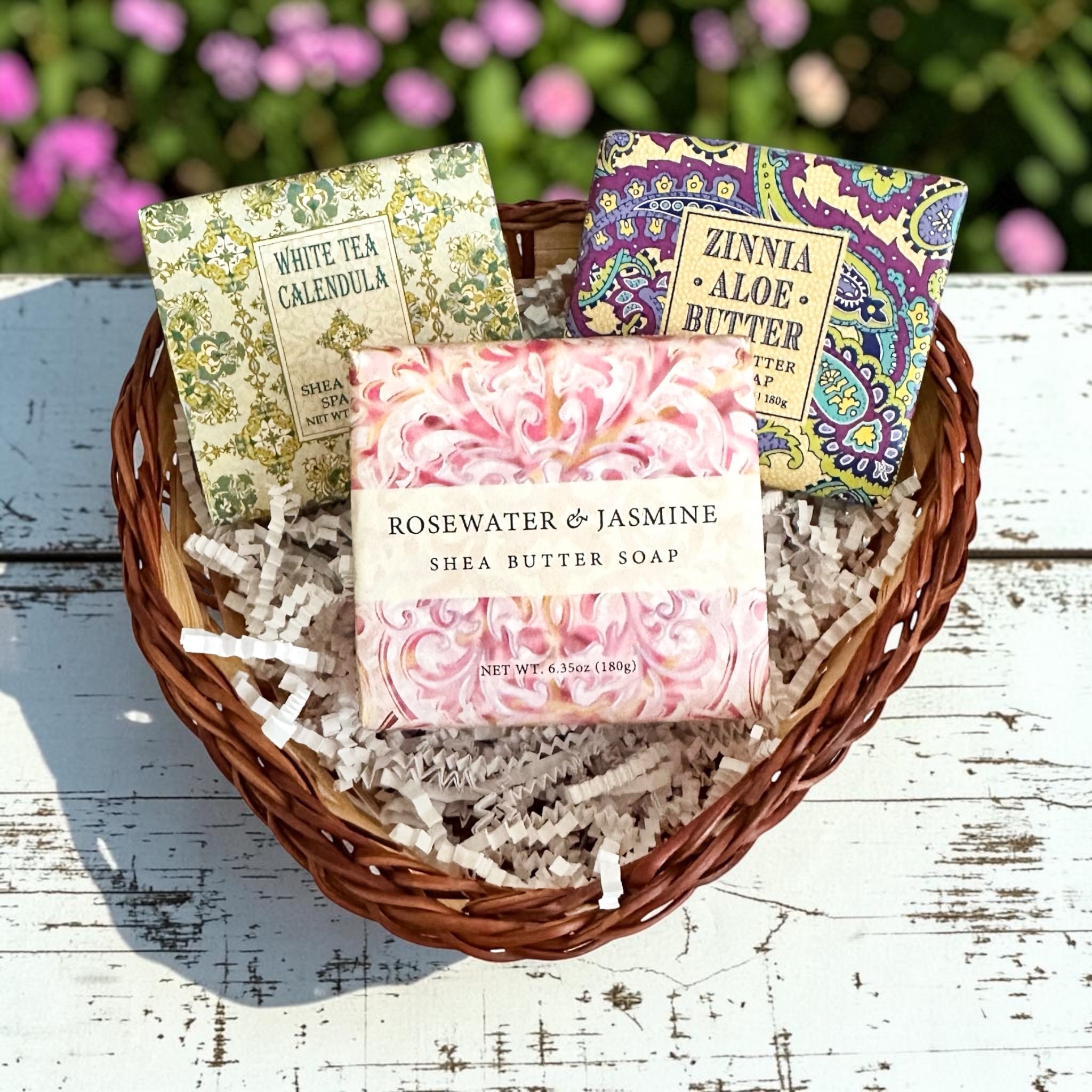 Three packaged shea butter bars arranged in a heart-shaped wicker basket with shredded paper, set on a white wooden surface with blurred pink flowers in the background. The center bar reads 'ROSEWATER & JASMINE SHEA BUTTER,' and the side bars read 'WHITE TEA CALENDULA' and 'ZINNIA ALOE BUTTER.
