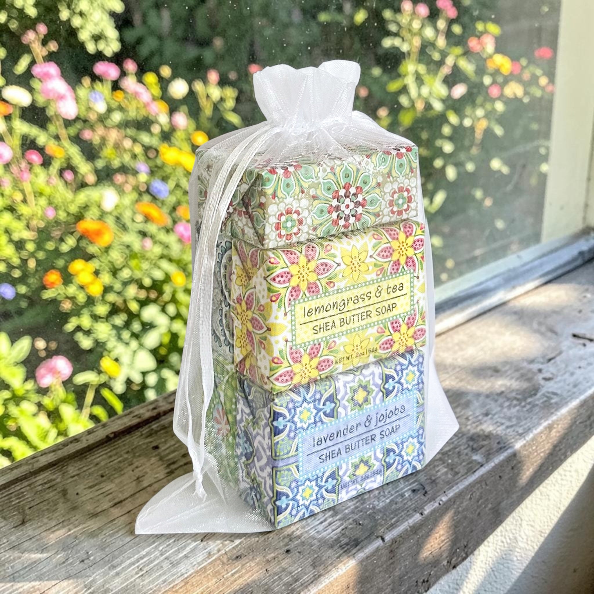 Five stacked bars of Shea Butter Soap in a clear drawstring bag, placed on a wooden windowsill with a blurred garden background. The soap bars feature colorful floral and geometric patterns and are labeled 'lemongrass & tea', 'shea butter soap', and 'lavender & lemon'.