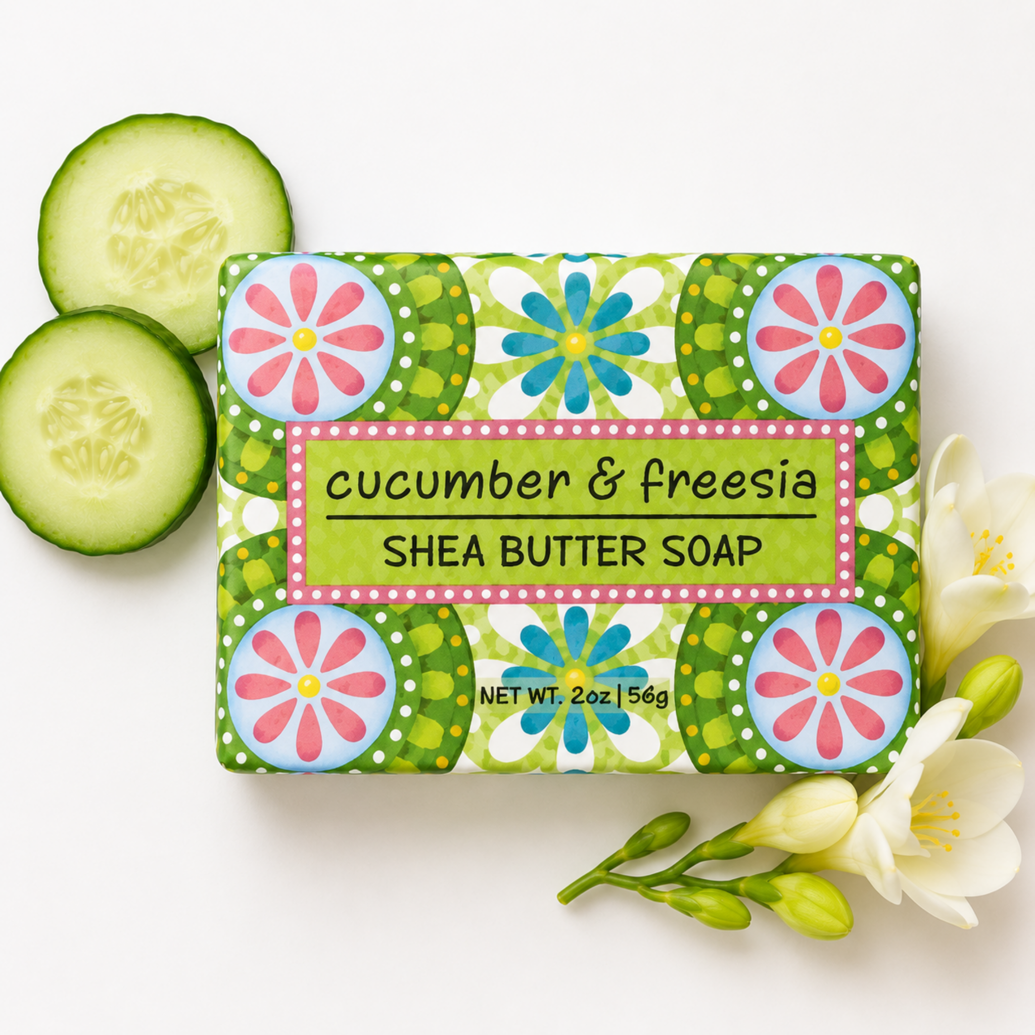 A rectangular bar of shea butter soap wrapped in a colorful floral-patterned paper sleeve, labeled 'cucumber & freesia SHEA BUTTER SOAP,' arranged on a white background with cucumber slices, white flowers, and green buds.