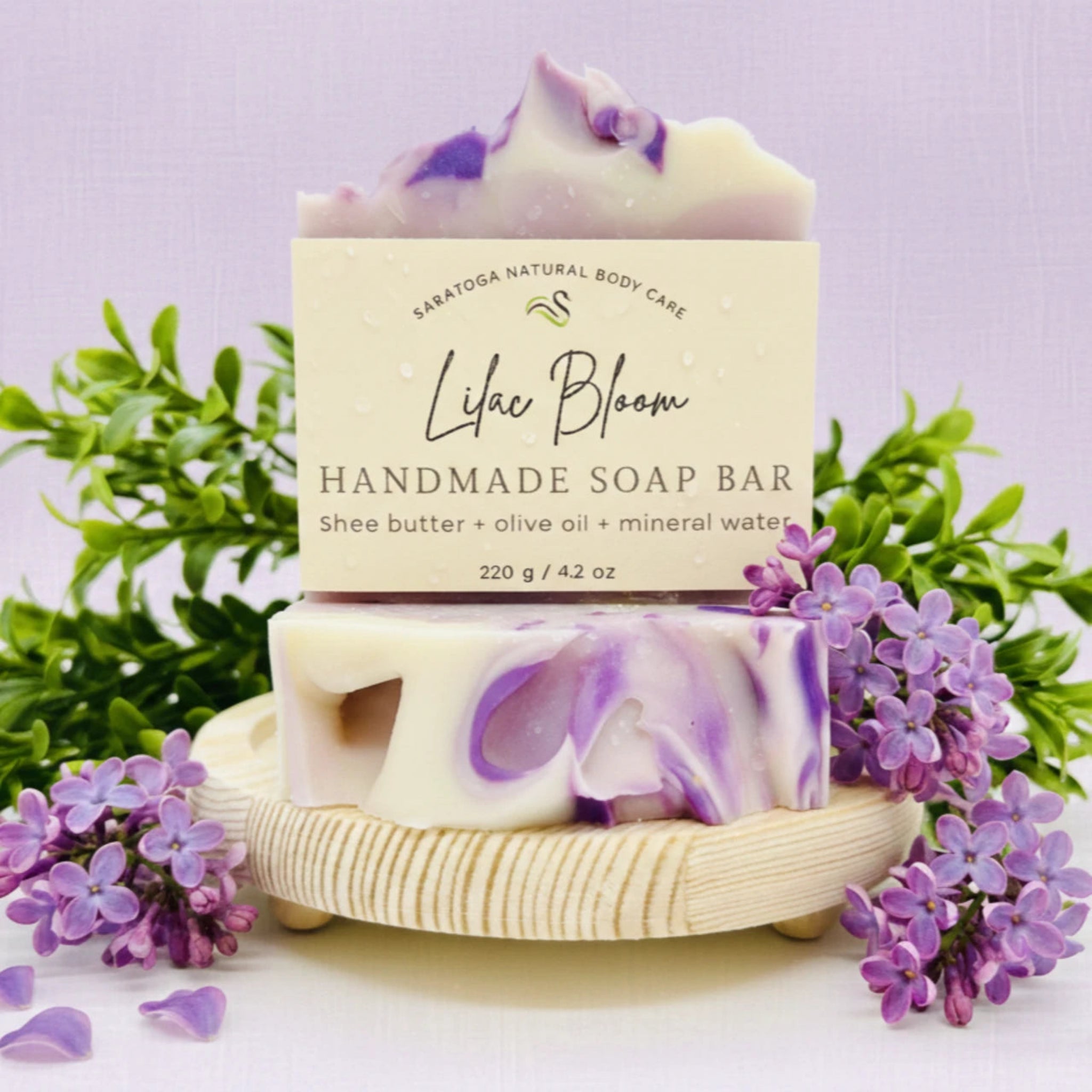 A handmade soap bar labeled 'Like Bloom' by Saratoga Natural Body Care, styled with purple flowers and green leaves on a light wooden dish against a soft lavender background.