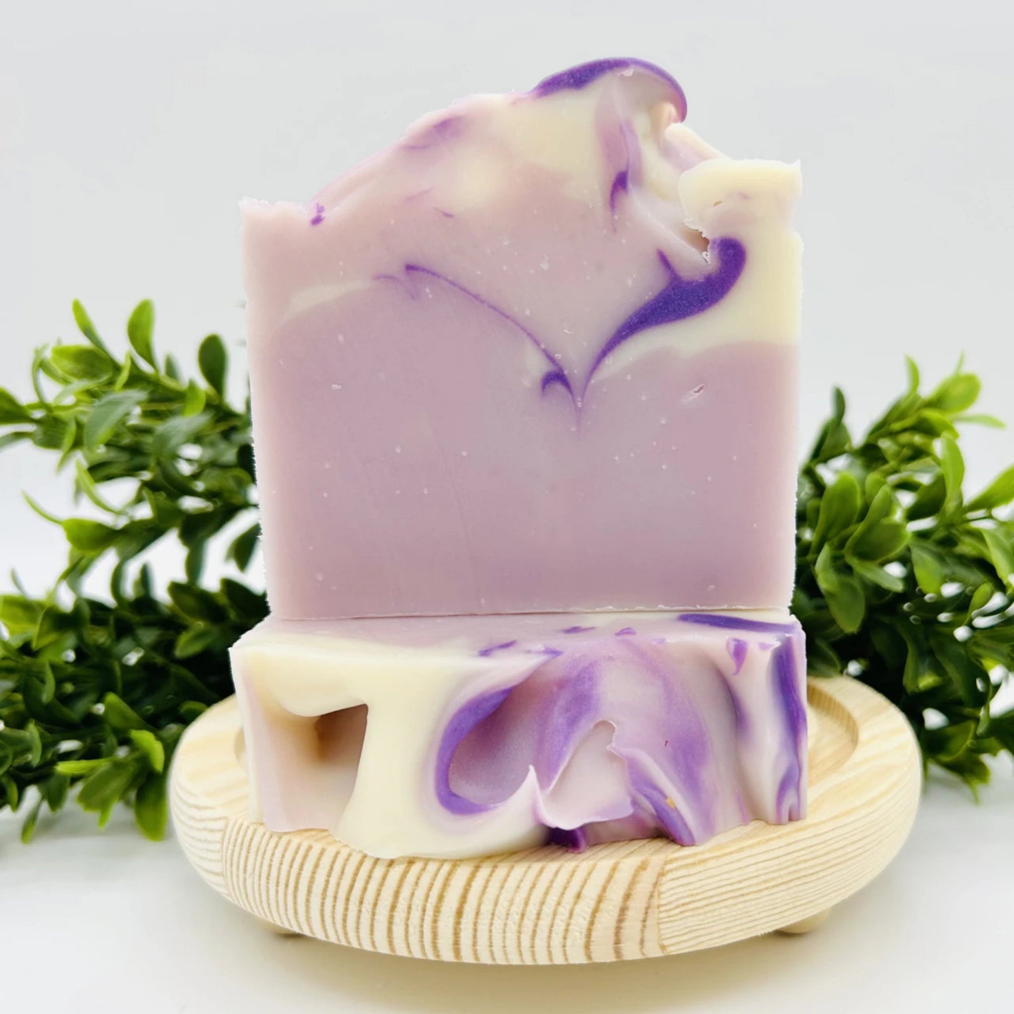 A handmade lavender and cream soap bar with purple swirls, displayed on a small wooden soap dish with green foliage in the background.