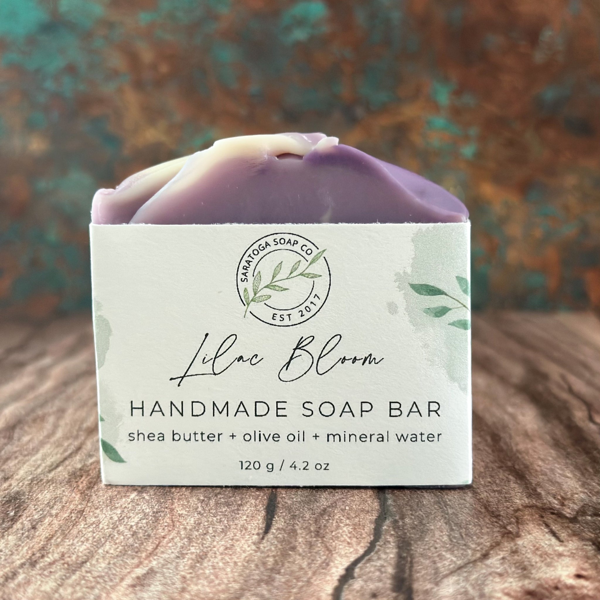 A handmade soap bar wrapped in a white paper label reading 'Lilac Bloom Handmade Soap Bar' by Sanctuary Soap Co, resting on a rustic wooden surface with a blurred teal and brown background.