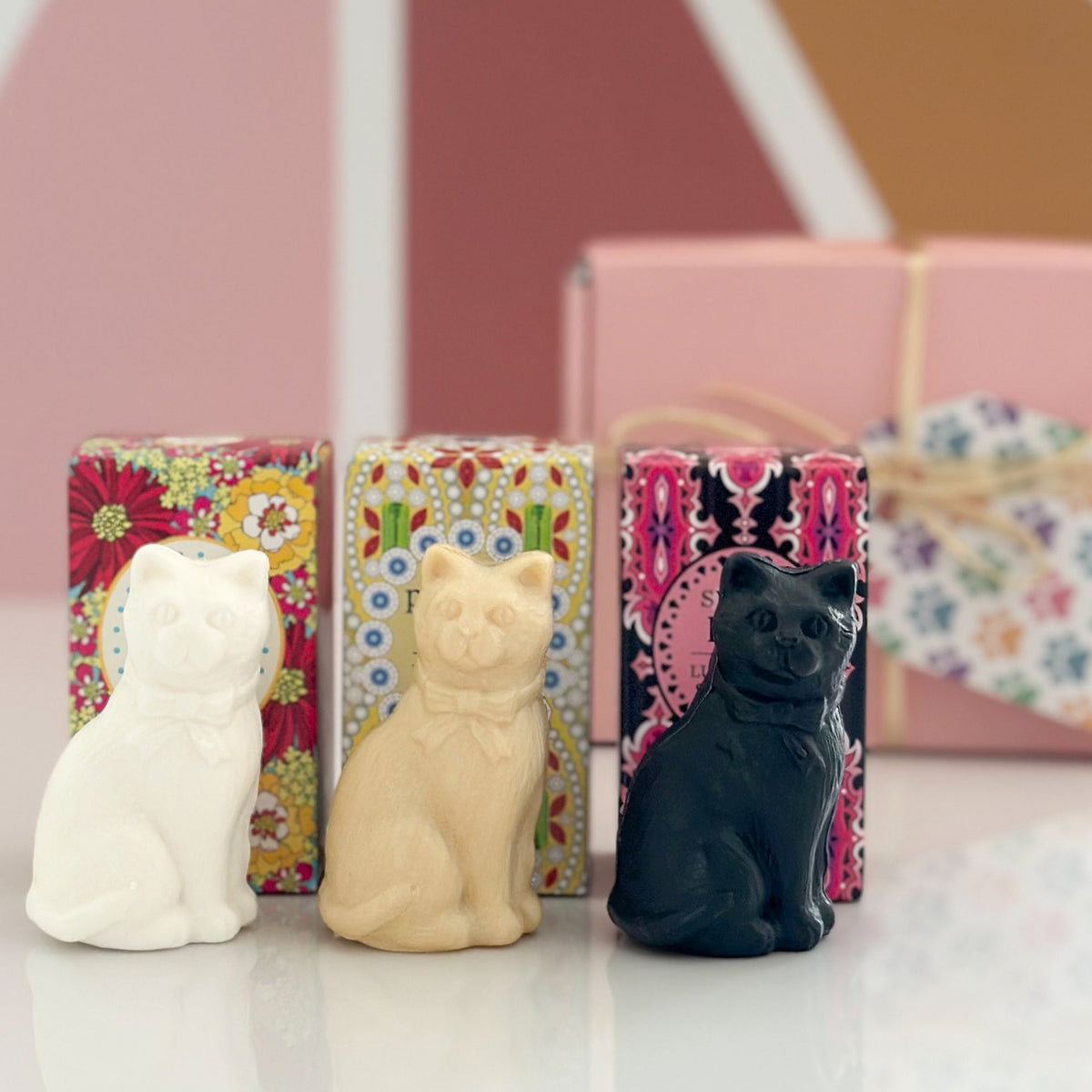 Three small cat-shaped soap bars in white, cream, and black, displayed in front of their colorful patterned gift boxes on a white surface.