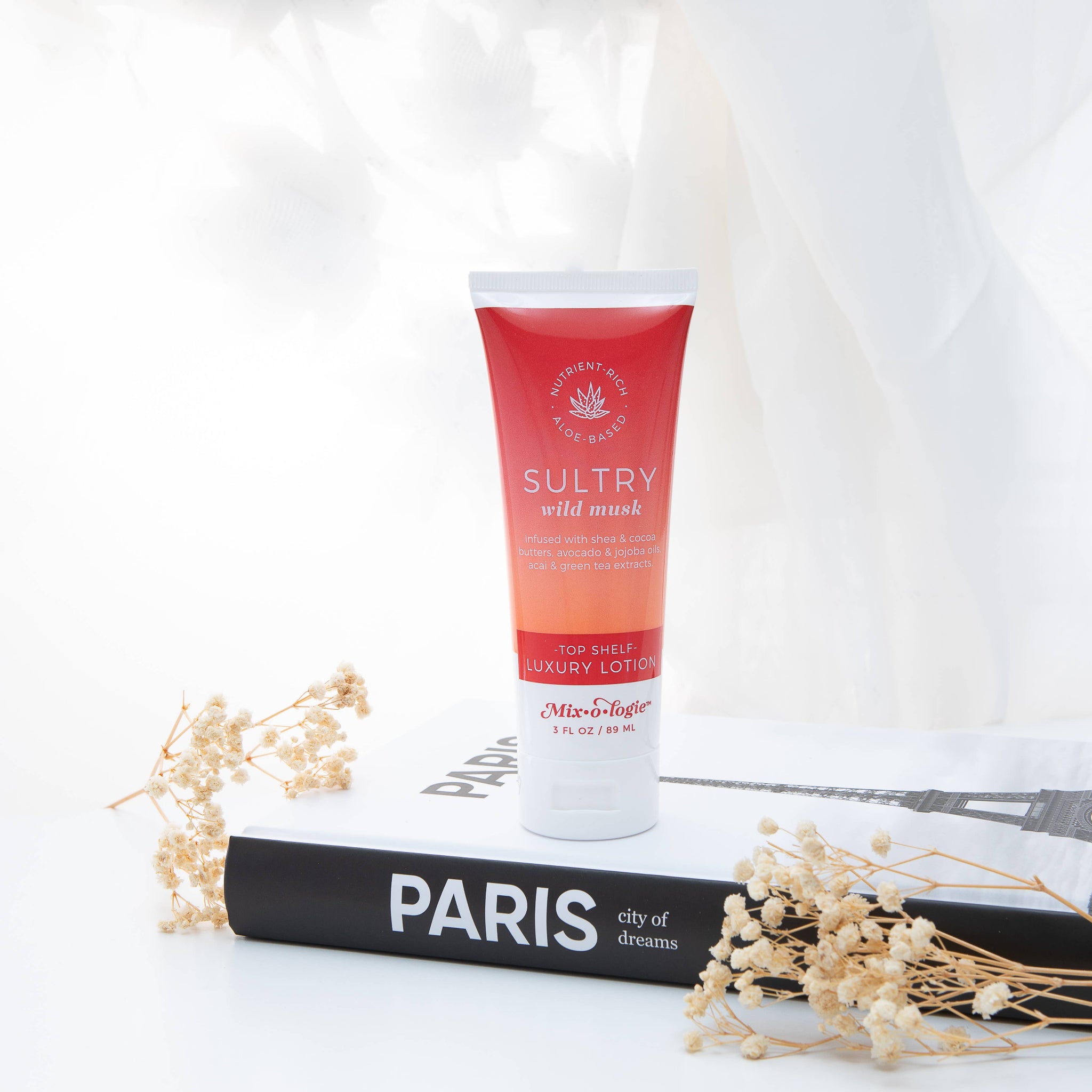 A squeeze tube of Sultry Wild Mask luxury lotion by MisEuphoria, standing upright on a black book with dried flowers around it against a white draped background.