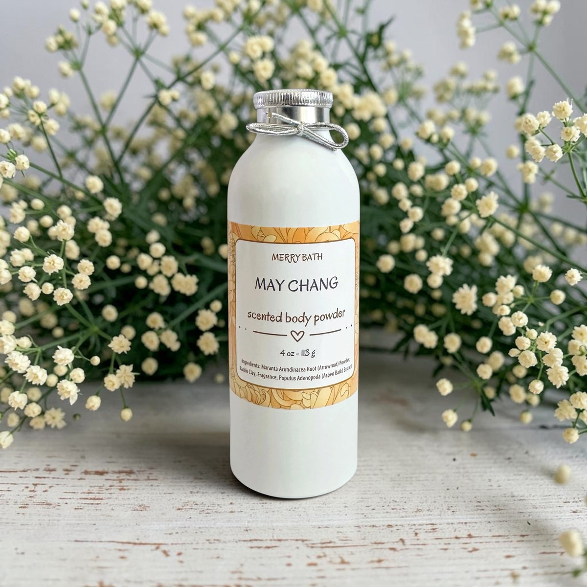 A white bottle of Merry Bath May Chang scented body powder with a silver metal cap and a decorative label, styled on a rustic white wooden surface with white baby's breath flowers in the background.