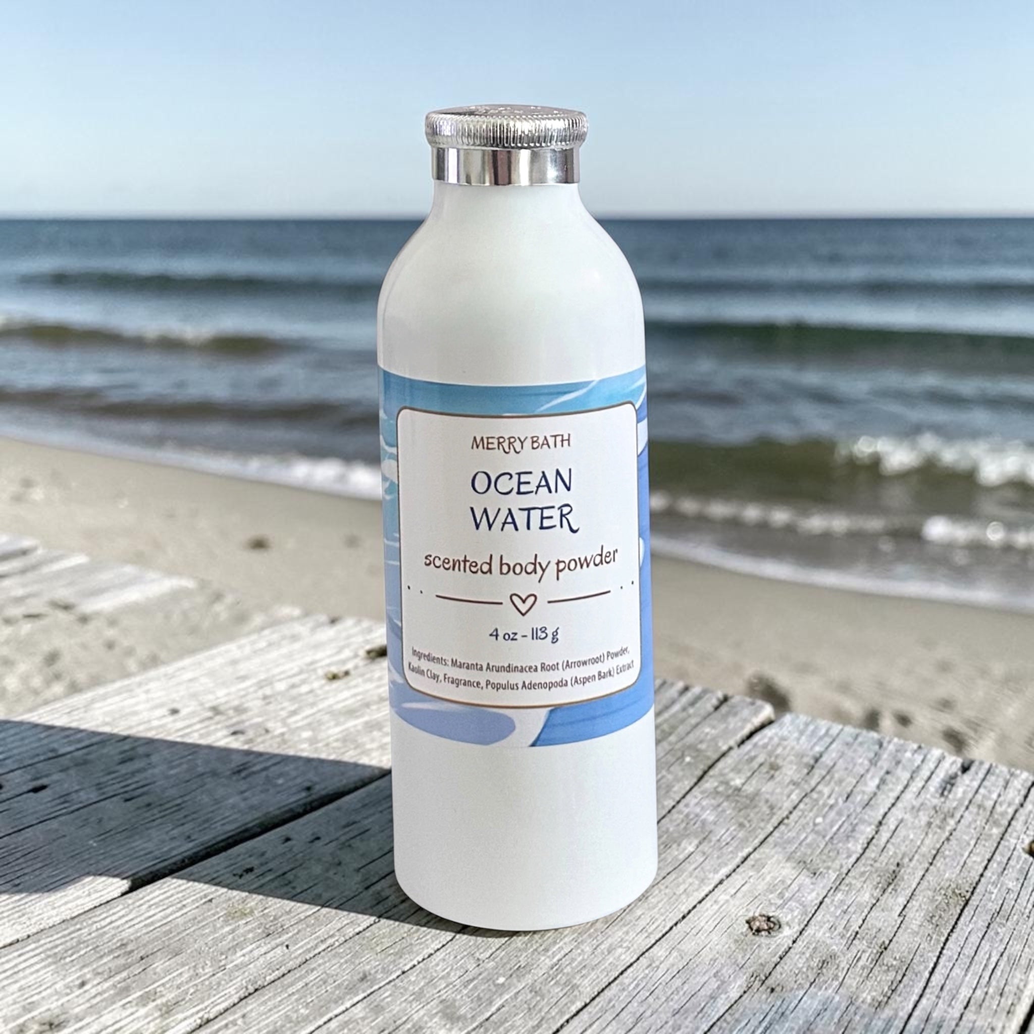 A white bottle of Merry Bath Ocean Water scented body powder with blue accents, placed on a weathered wooden board with the ocean and beach in the background.