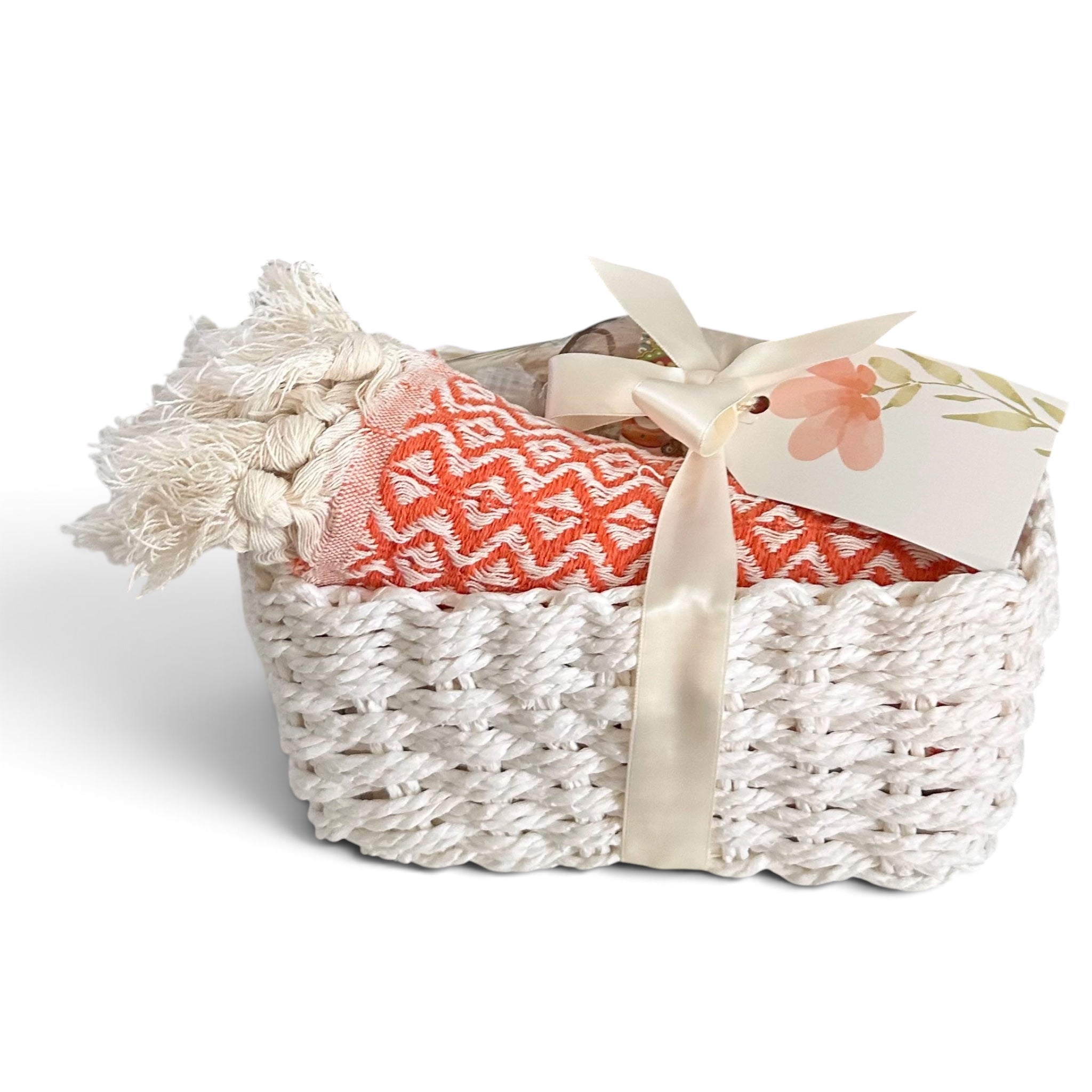 A white woven basket containing a folded coral-orange and white patterned throw blanket with a cream tassel, tied with a cream satin ribbon and a small floral gift tag, on a white background.