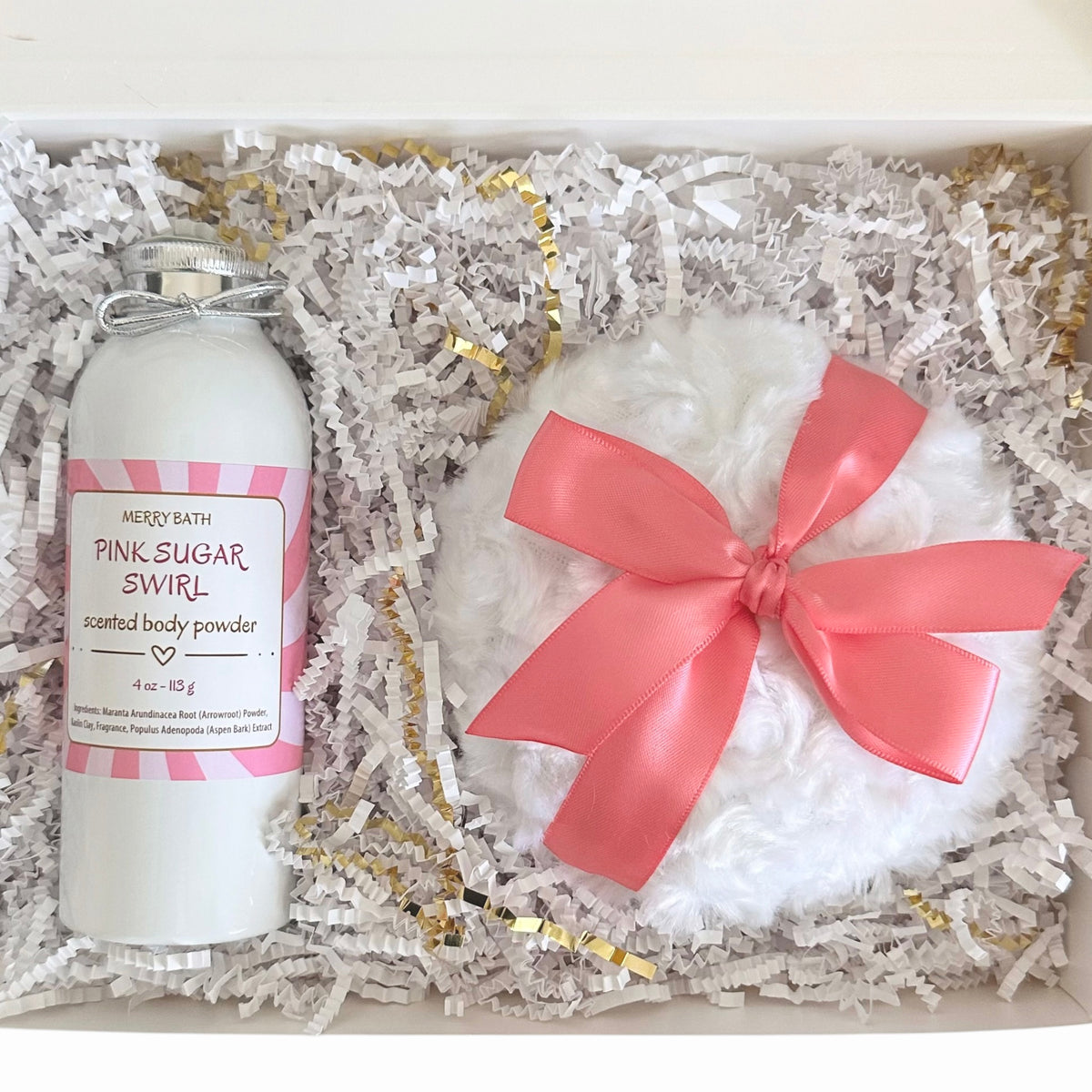 A gift set in a white box filled with white shredded paper and gold tinsel, featuring a white bottle labeled Merry Bath Pink Sugar Swirl' scented body powder and a white fluffy bath bomb with a large pink ribbon bow.
