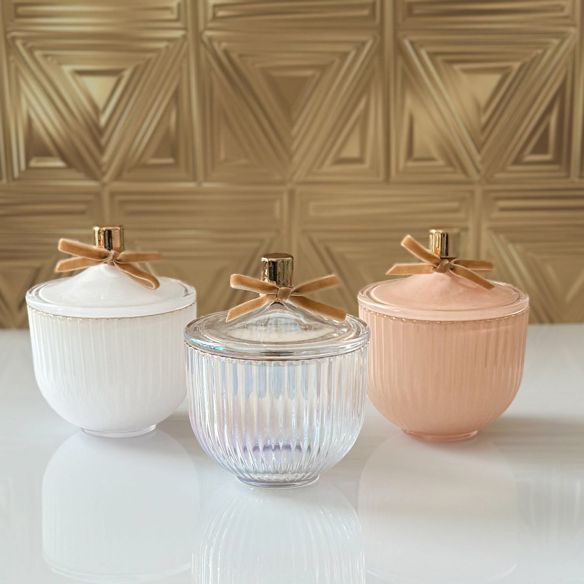 Three ribbed lidded jars with gold-tone tops and ribbon bows, shown in white, clear, and blush pink on a glossy white surface against a metallic gold wall.
