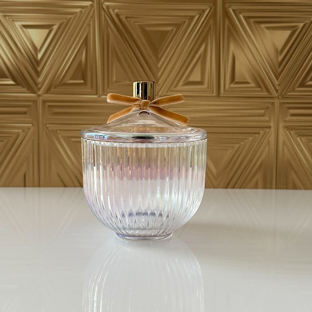 A clear ribbed glass jar with a matching glass lid, topped with a small gold cap and a tan ribbon bow, set on a white reflective surface against a gold geometric wall.