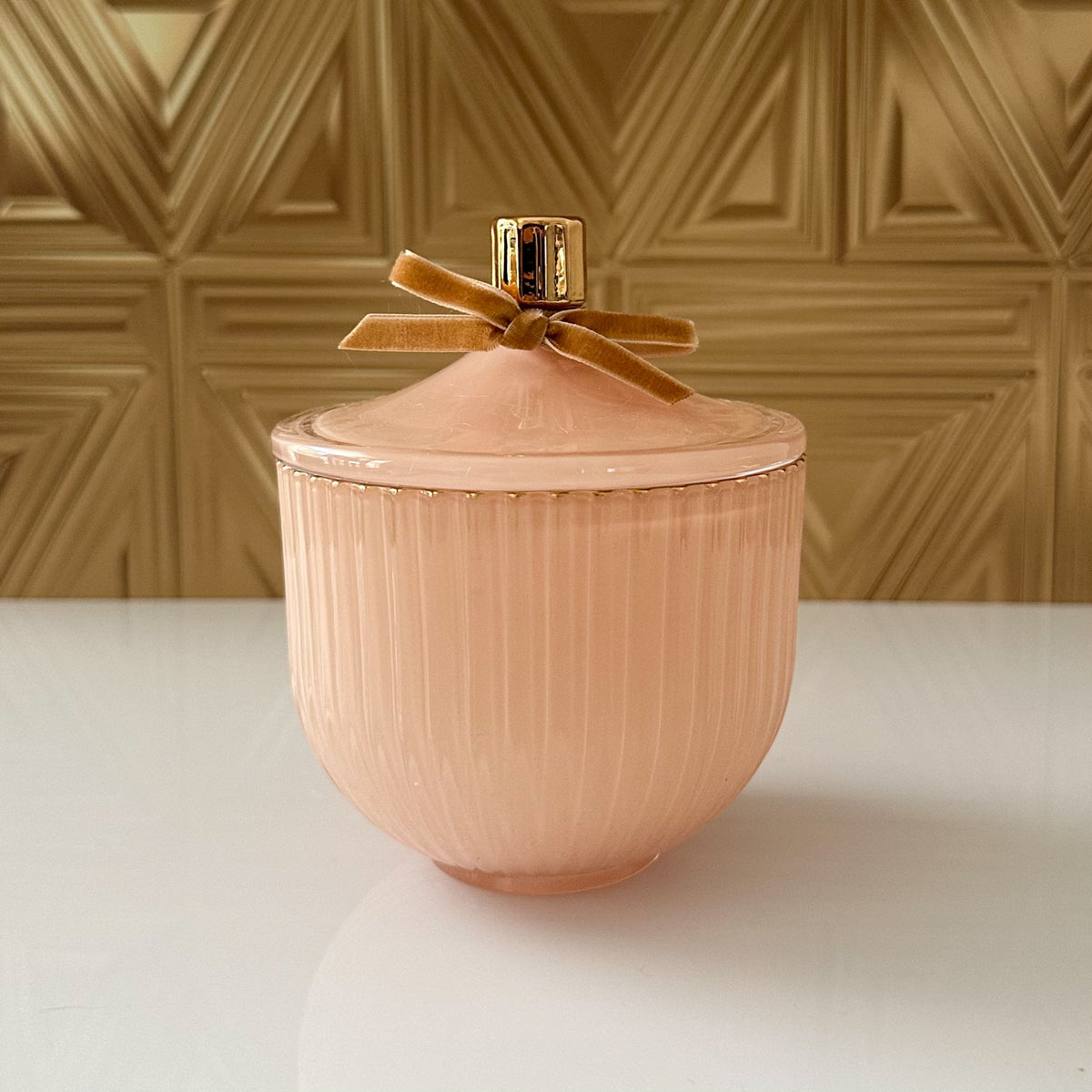 A ribbed dusty pink ceramic jar with a matching lid, decorated with a gold-tone handle and a velvet ribbon bow, set against a gold geometric wall background.