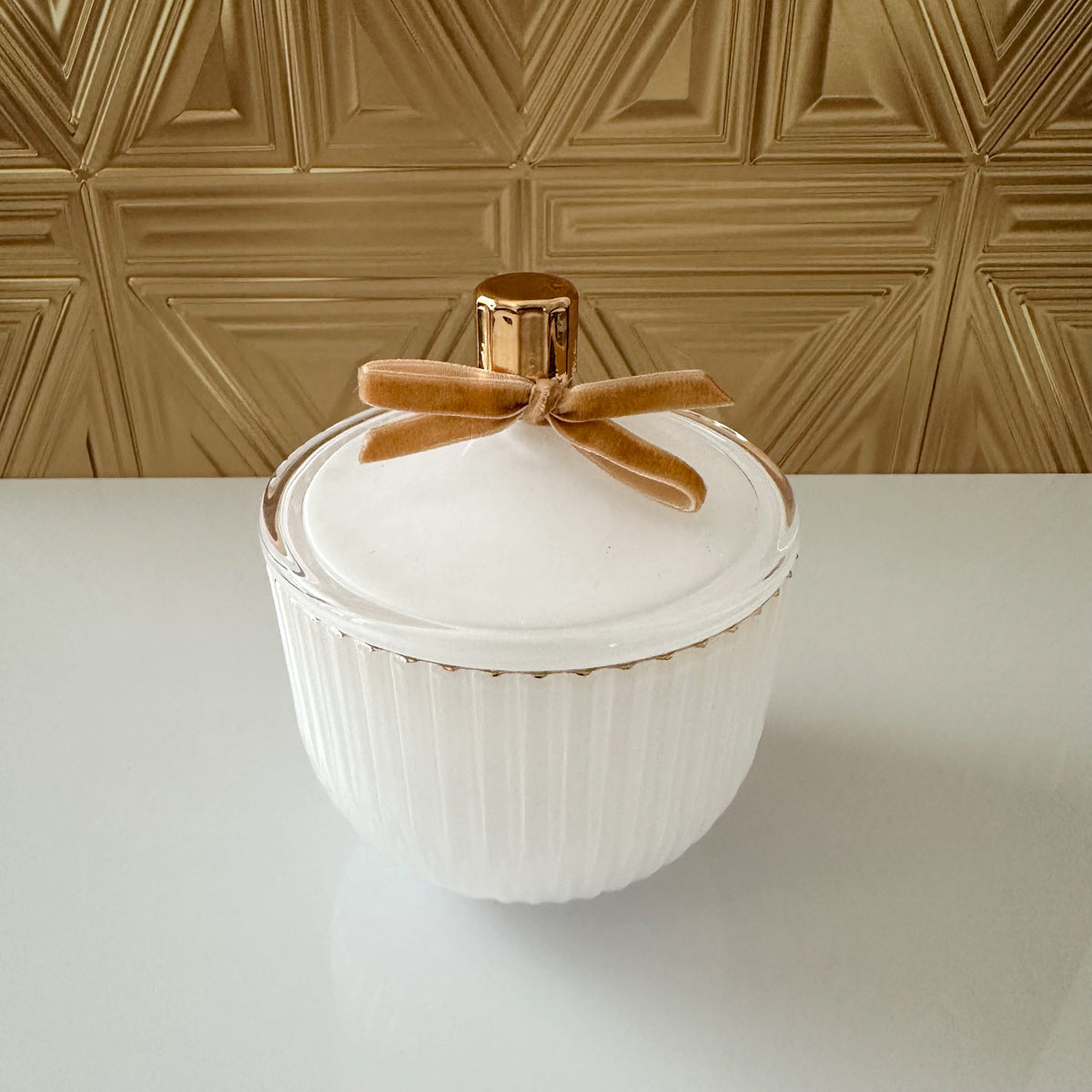 A white ribbed ceramic lidded container with a metallic gold cap and a brown velvet ribbon bow, set on a white surface against a gold geometric wall.