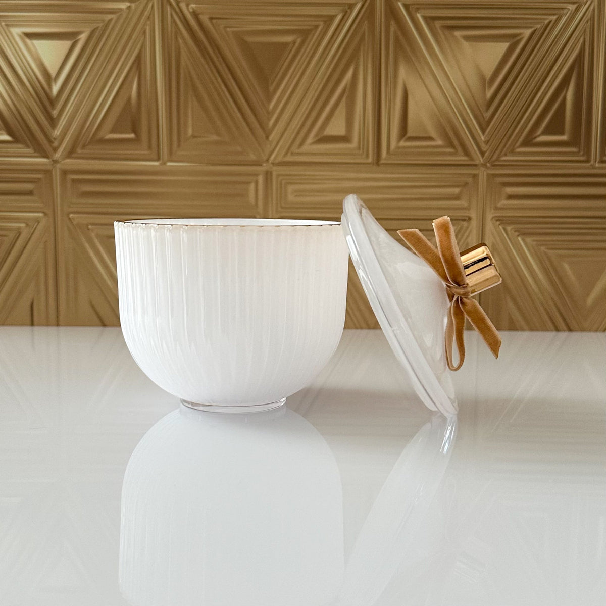 A white ribbed ceramic lidded container with a clear glass lid tied in a gold ribbon, set on a glossy white surface against a textured gold geometric wall.