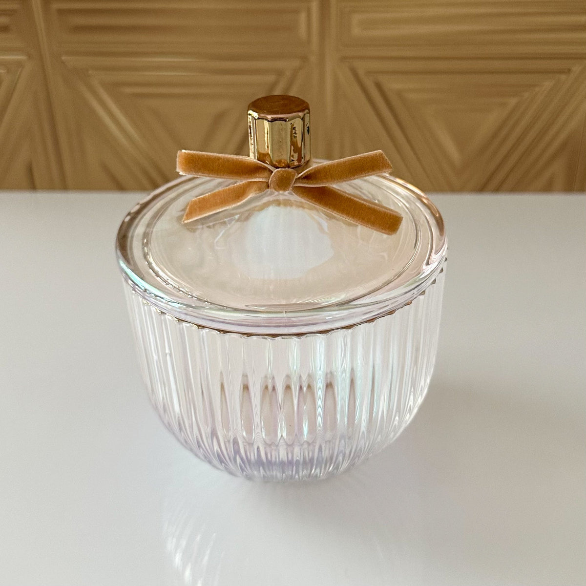 A clear ribbed glass jar with a matching glass lid, topped with a gold cap and a tan velvet bow, set on a white surface against a beige geometric wall.