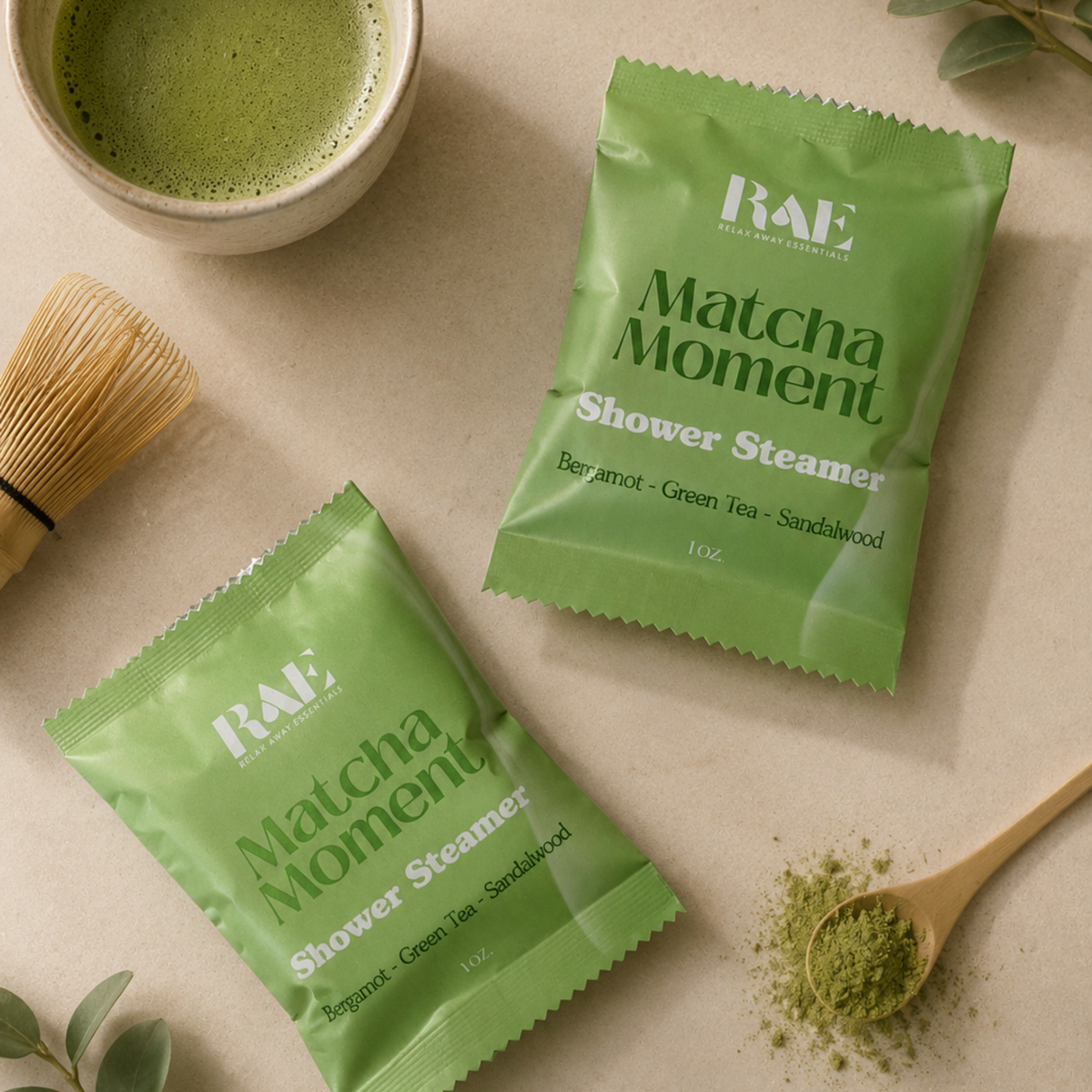 Two green RAE Matcha Moment Shower Steamer packets arranged on a beige surface with a bowl of green tea, a bamboo whisk, a wooden spoon with matcha powder, and green leaves.