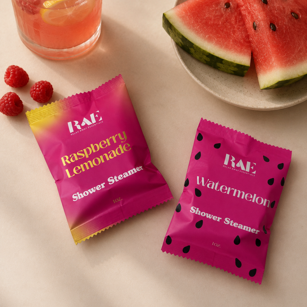 Two pink shower steamer packets labeled 'Raspberry Lemonade' and 'Watermelon' next to a glass of raspberry lemonade and watermelon slices.