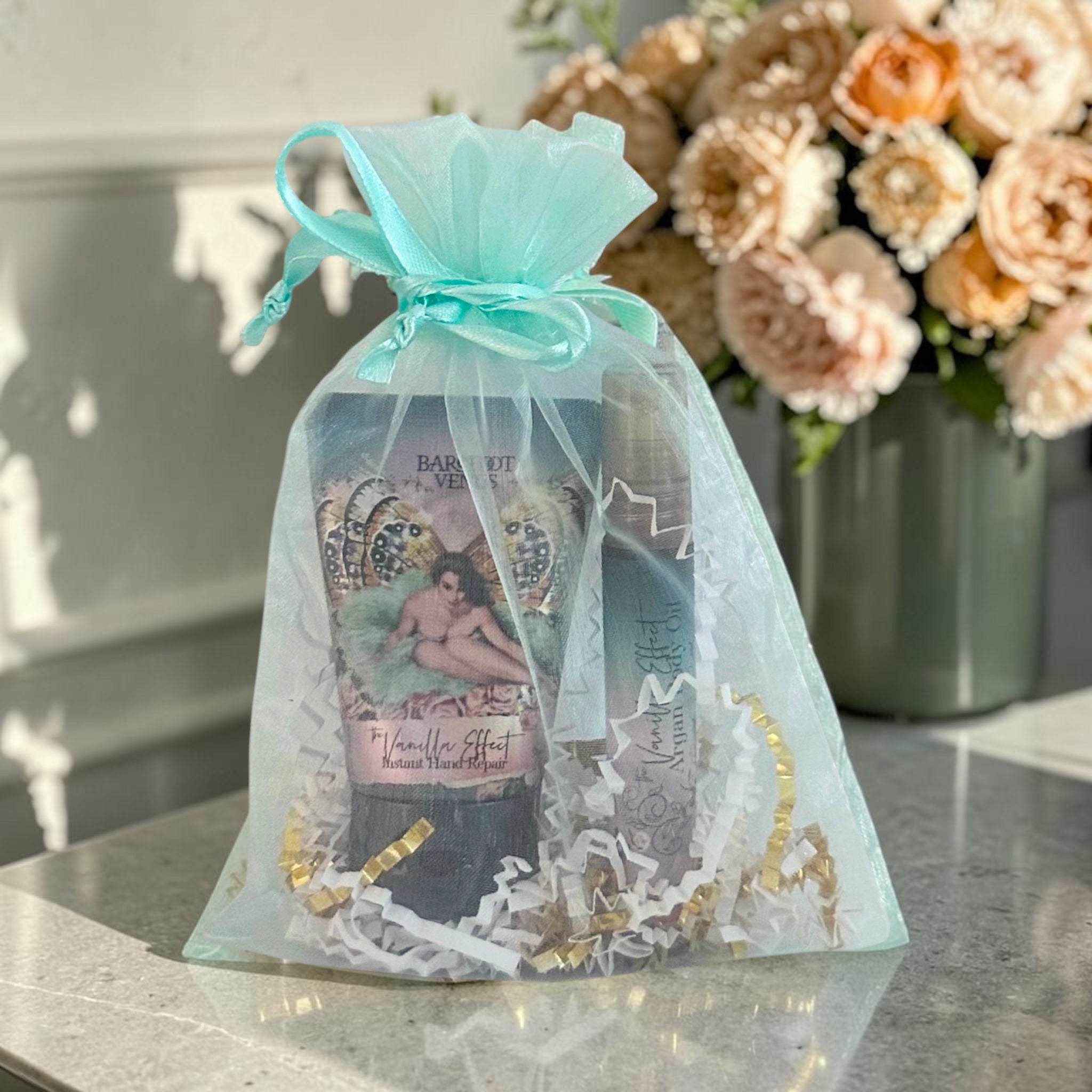 A gift set in a sheer light blue organza bag tied with a matching ribbon, placed on a marble surface with a vase of flowers in the background. The set includes a hand cream tube labeled 'Barsettes' and a boxed item labeled 'Manicure Effect'.