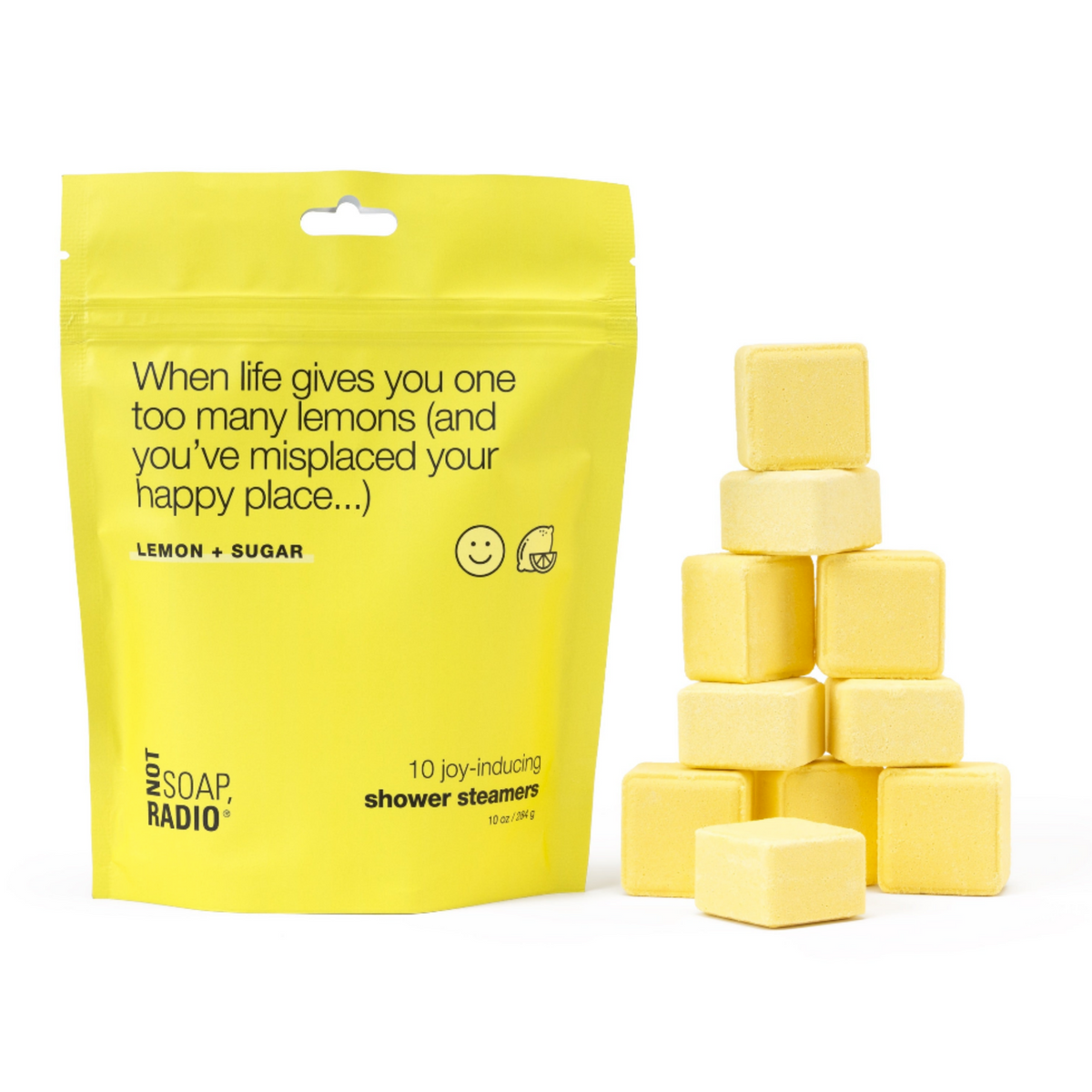 A bright yellow stand-up pouch labeled 'Lemon + Sugar' and '10 joy-Inducing shower steamers' stands next to a small stack of pale yellow cube-shaped shower steamers on a white background.