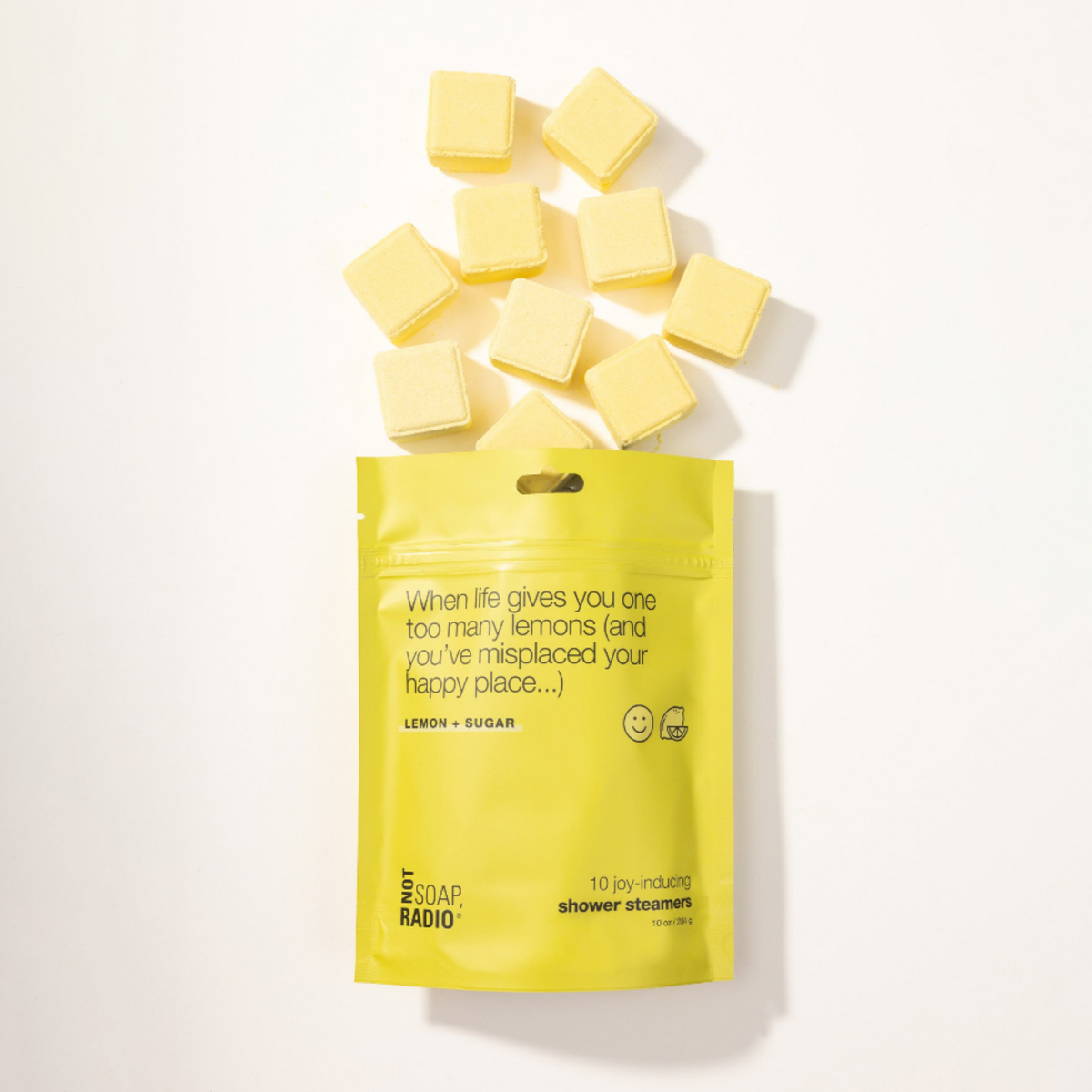 A bright yellow stand-up pouch labeled 'The Soap Radio' with the scent name 'Lemon + Sugar,' shown on a white background with several pale yellow cube-shaped shower steamers scattered above it.