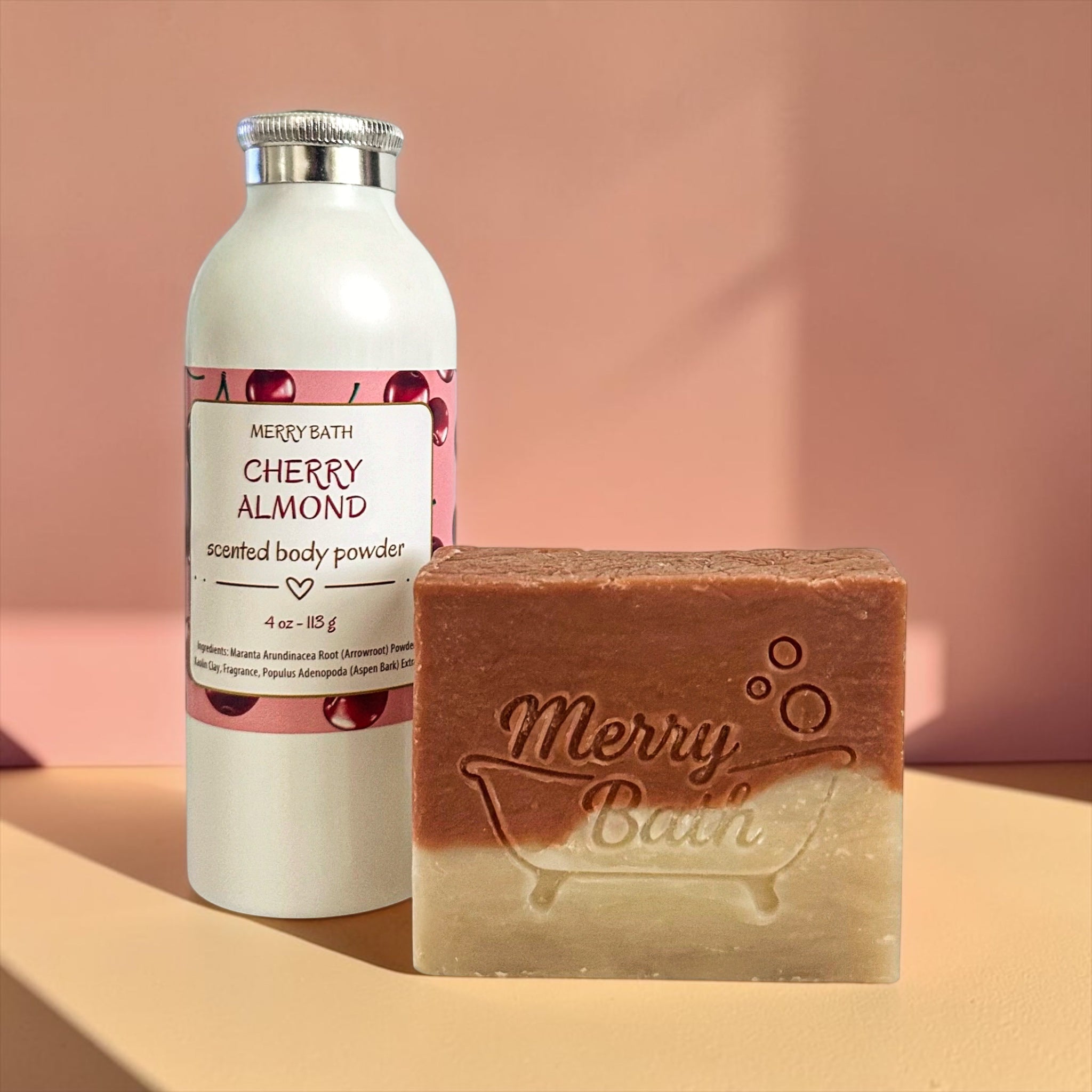 Bottle of cherry almond scented body powder and a bar of soap with 'Merry Bath' branding on a peach background.