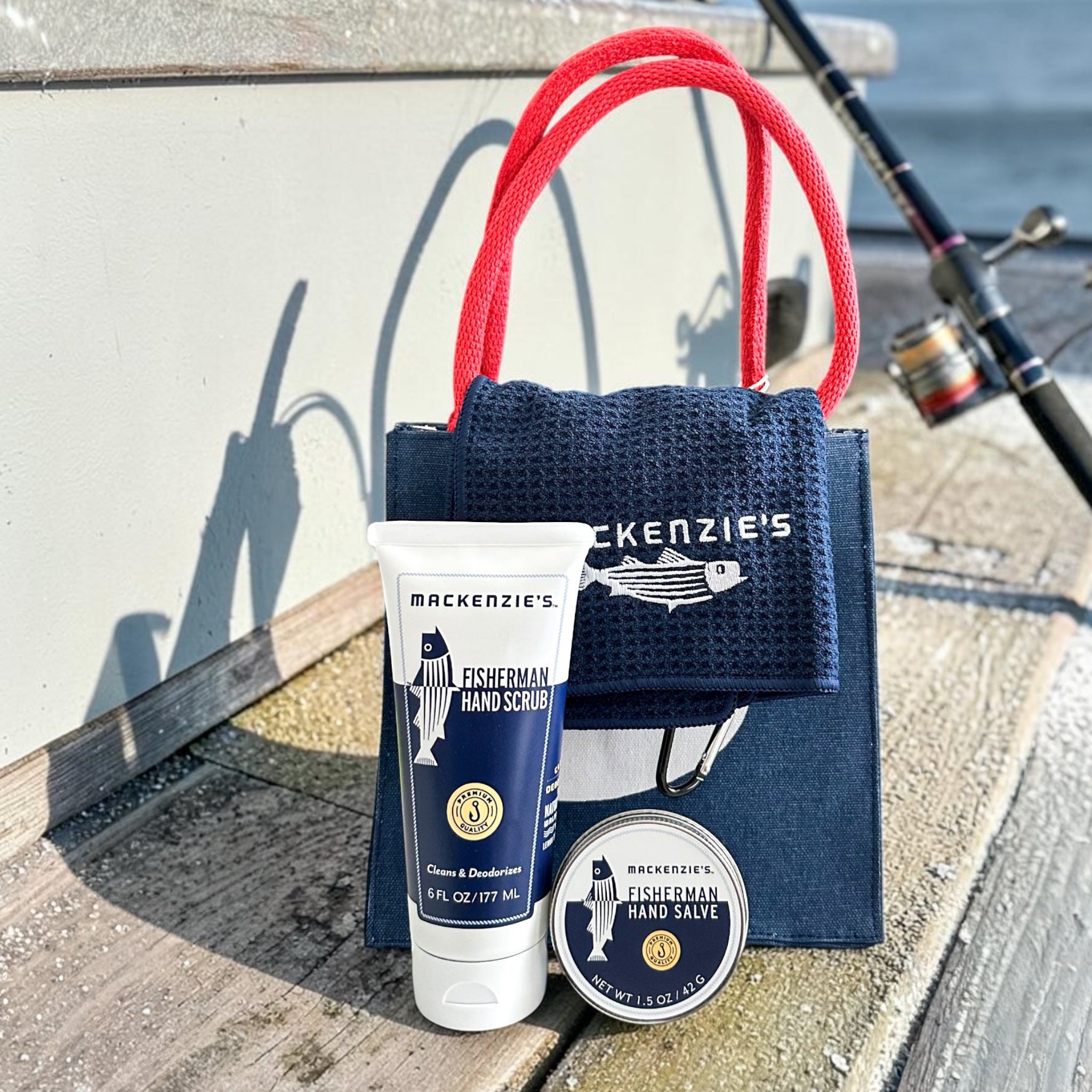 A fishing-themed gift set from Mackenzie's Hand Scrubs and Hand Salves, including a navy blue gift bag with red handles, a hand scrub tube, and a tin of hand salve, arranged on a wooden dock next to a fishing rod.