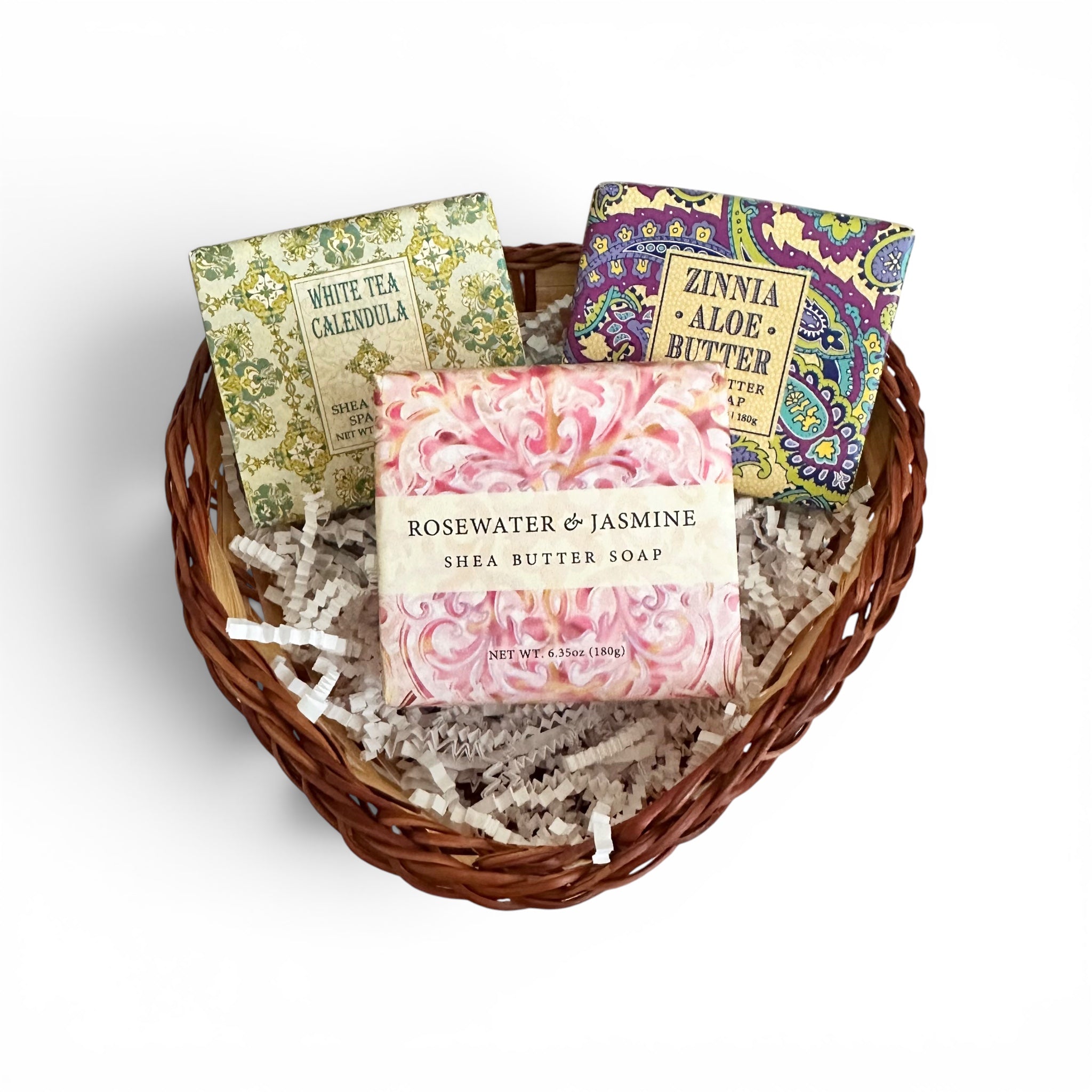 A heart-shaped wicker basket filled with shredded paper and three decorated soap bars. The soaps have floral and paisley patterns in green, purple, and pink, with labels reading 'White Tea Calendula', 'Zinnia Aloe Butter', and 'Rosewater & Jasmine Shea Butter Soap'.