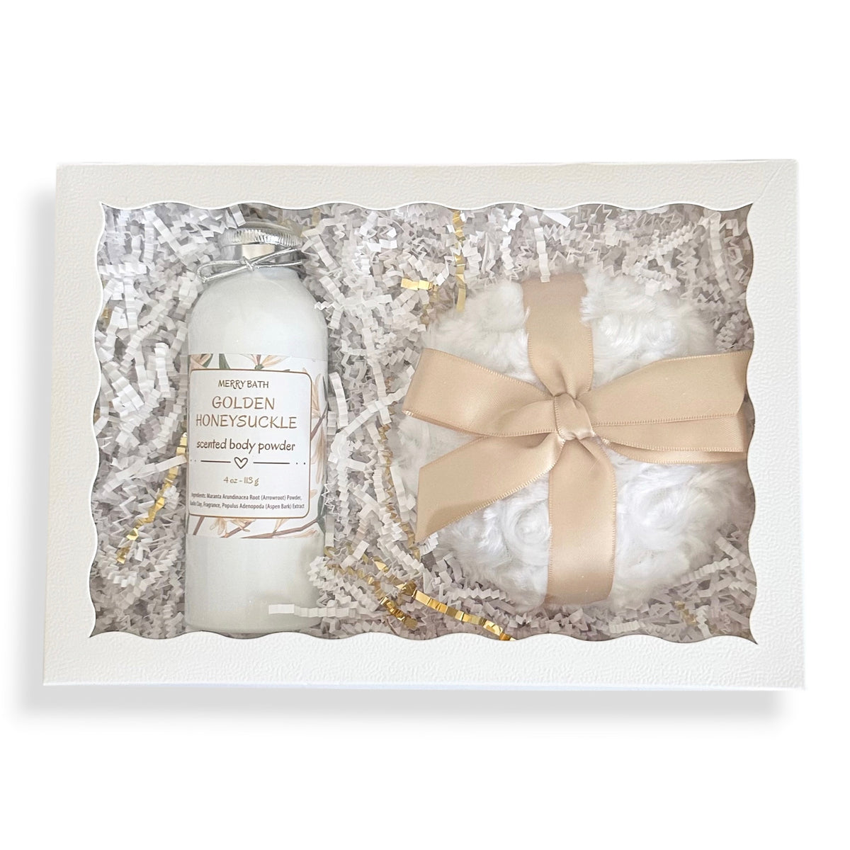 A gift set in a white box containing a white bottle of Merry Bath Golden Honeysuckle natural body powder and a white bath bomb tied with a beige ribbon, surrounded by shredded paper packaging.