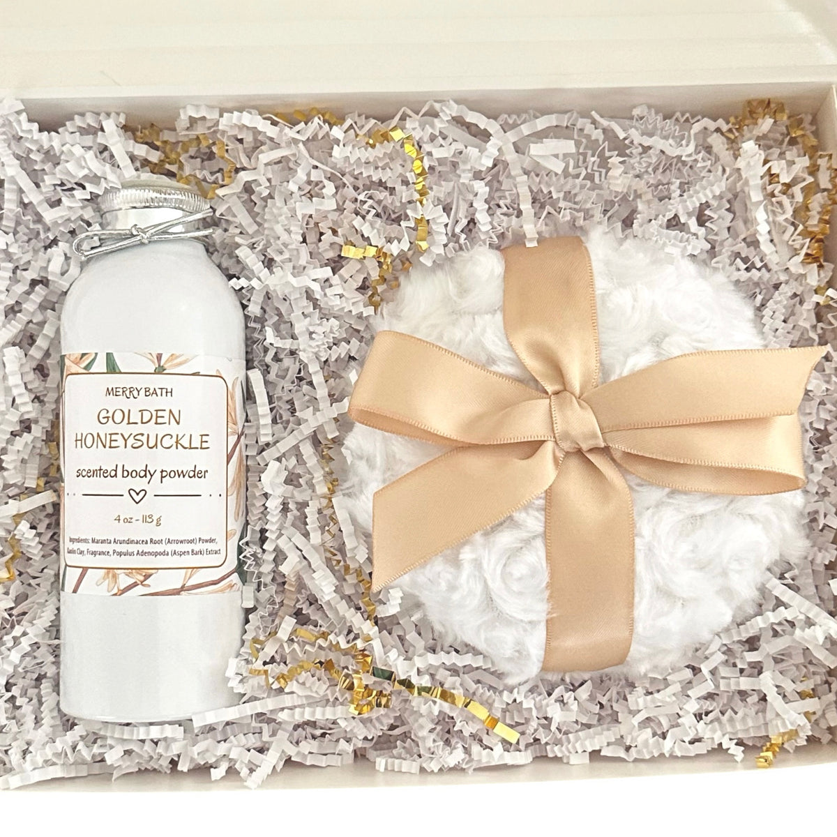 A gift set in a cream-colored box filled with white shredded paper, featuring a white glass bottle of Merry Bath Golden Honeysuckle scented body powder and a white fluffy bath bomb wrapped in a beige satin ribbon.