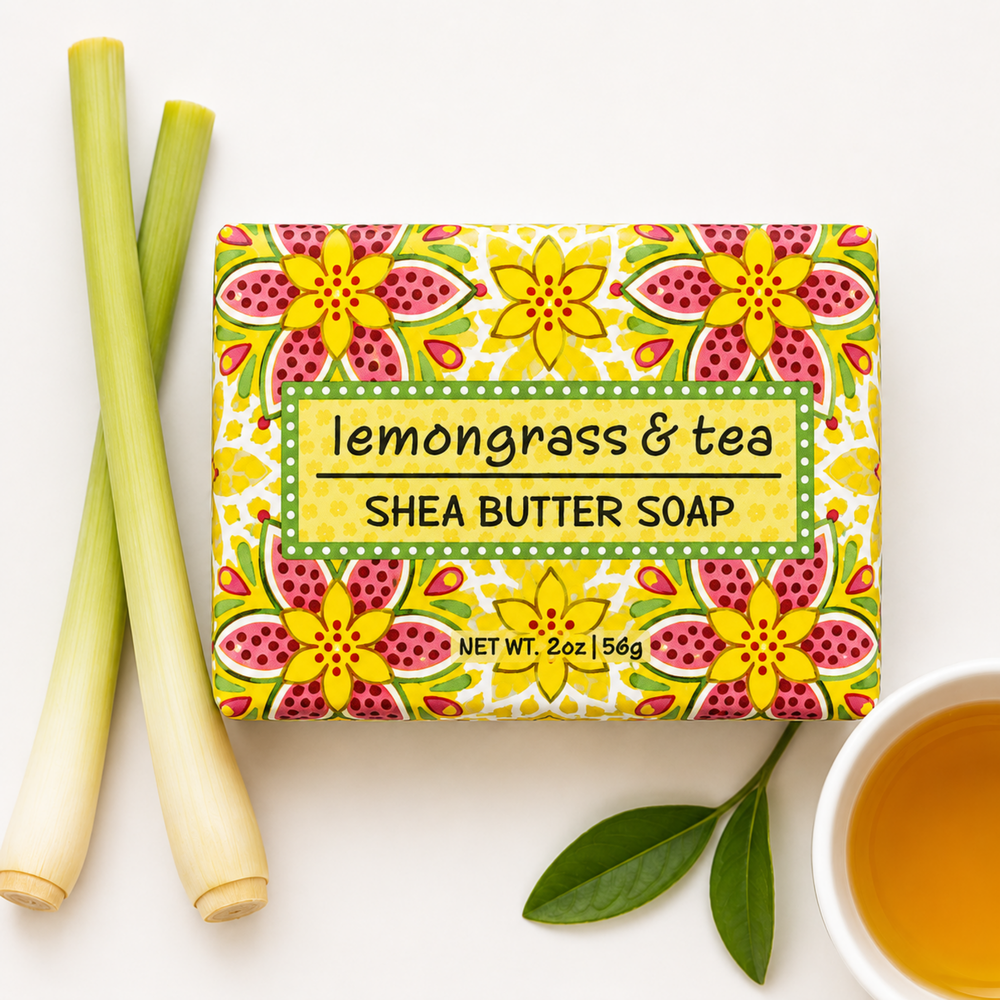 A rectangular bar of shea butter soap labeled 'lemongrass & tea' sits on a white background, styled with two lemongrass stalks, a green leaf, and a cup of tea. The soap packaging features a bright floral pattern in yellow, pink, and green.