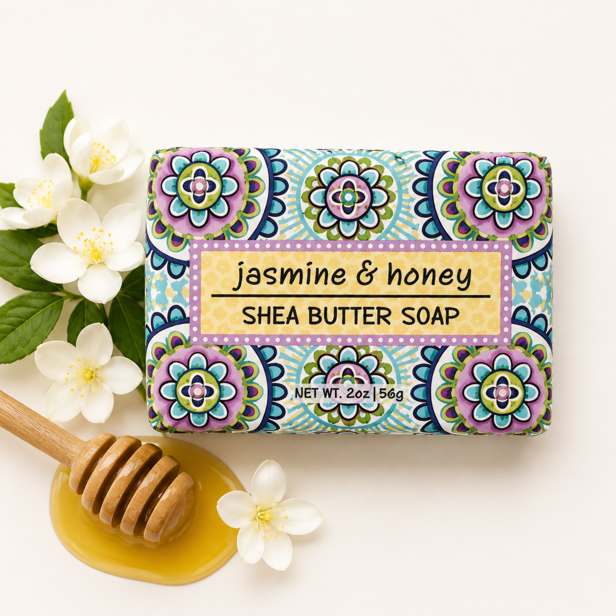 A rectangular bar of jasmine & honey shea butter soap with a colorful floral mandala pattern, styled with white jasmine flowers, a wooden honey dipper in honey, and a small white flower on a clean off-white background.