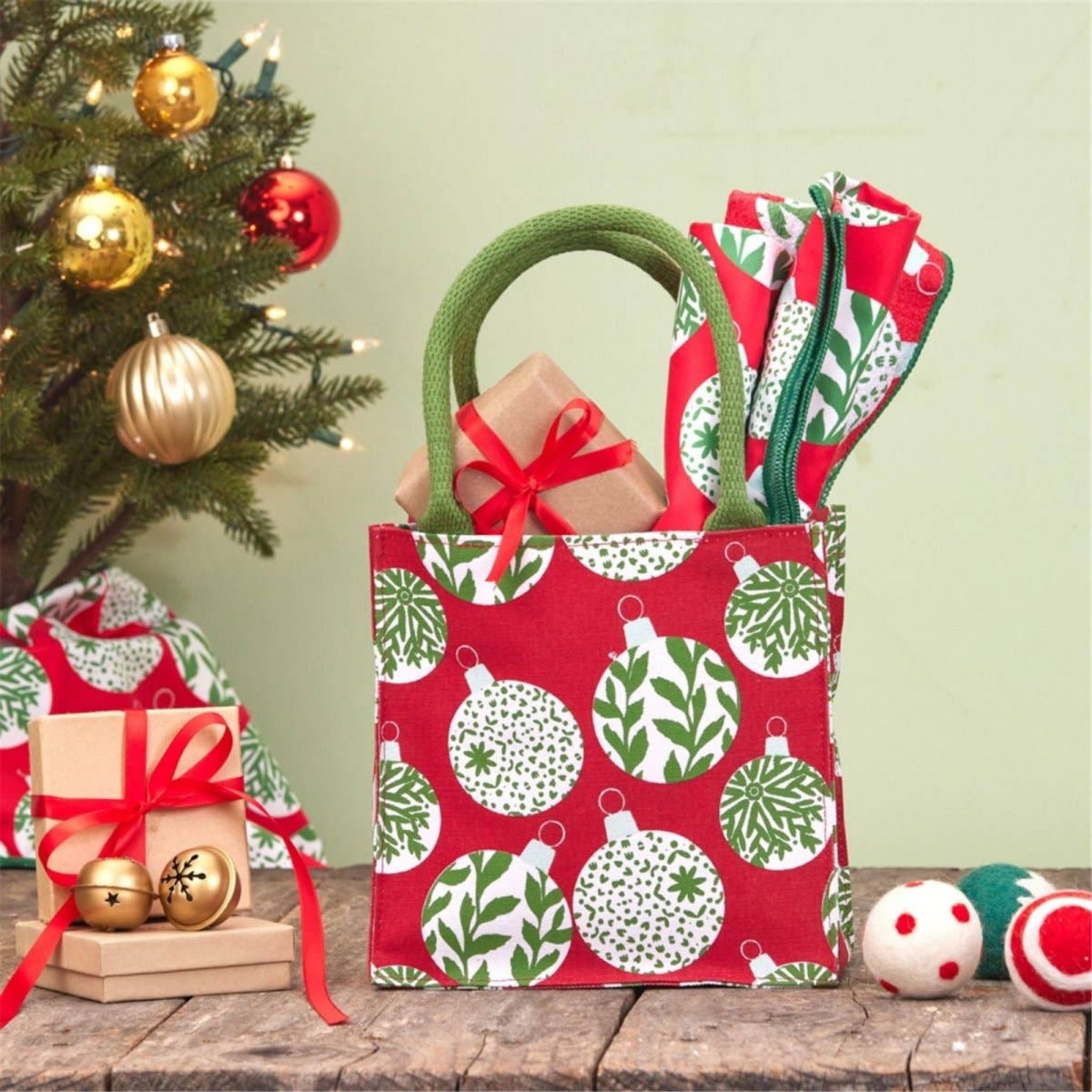 A red Christmas-themed gift bag with green handles and a white and green ornament pattern, filled with wrapped presents and standing on a rustic wooden surface next to a decorated Christmas tree.