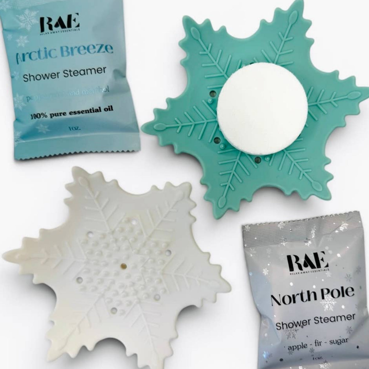 Two snowflake-shaped shower steamers, one teal and one white, shown with their respective RAE brand packets on a clean white background.