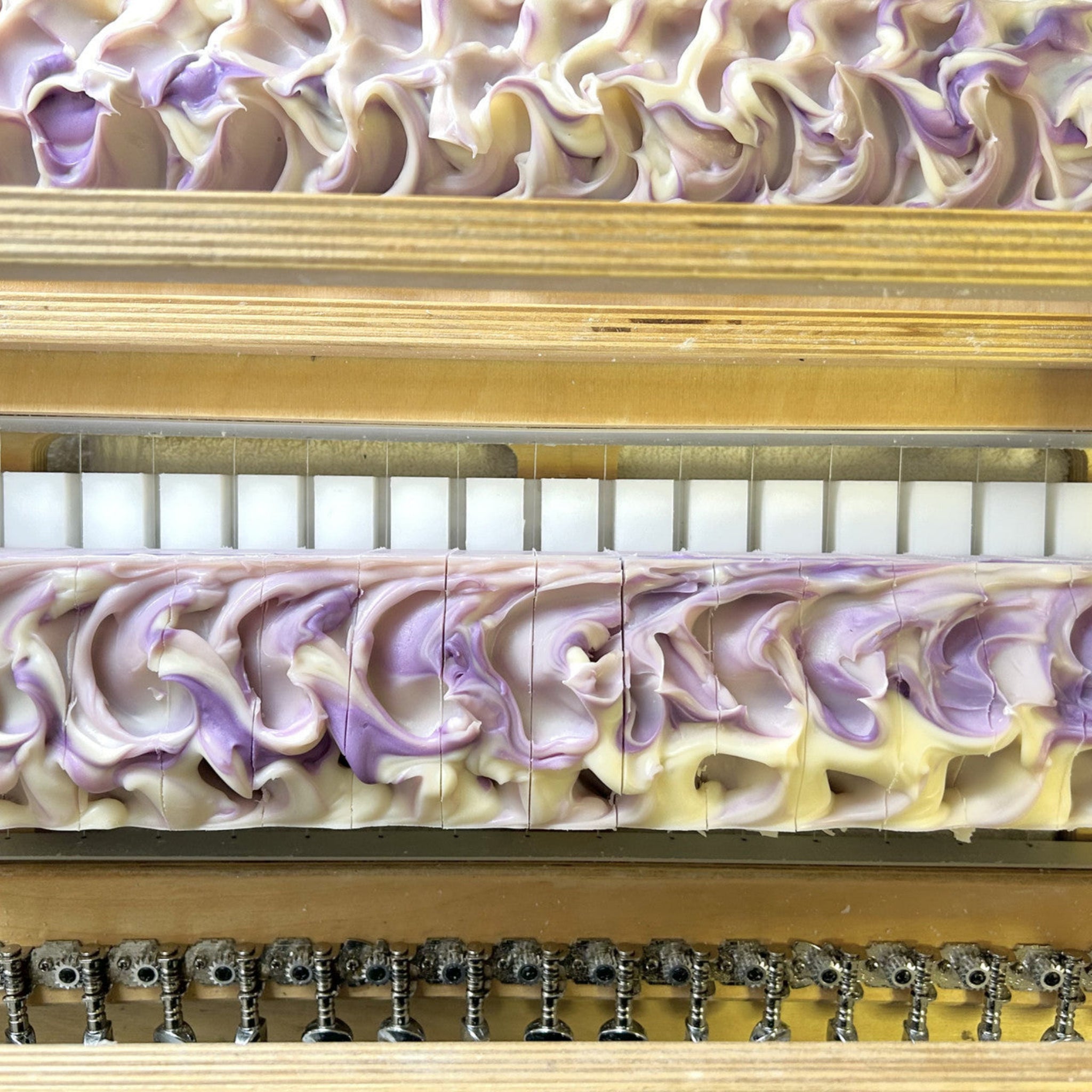 Close-up of handmade artisanal soap bars with a swirled lavender and cream pattern, arranged in a wooden soap mold with metal knobs visible along the bottom edge.