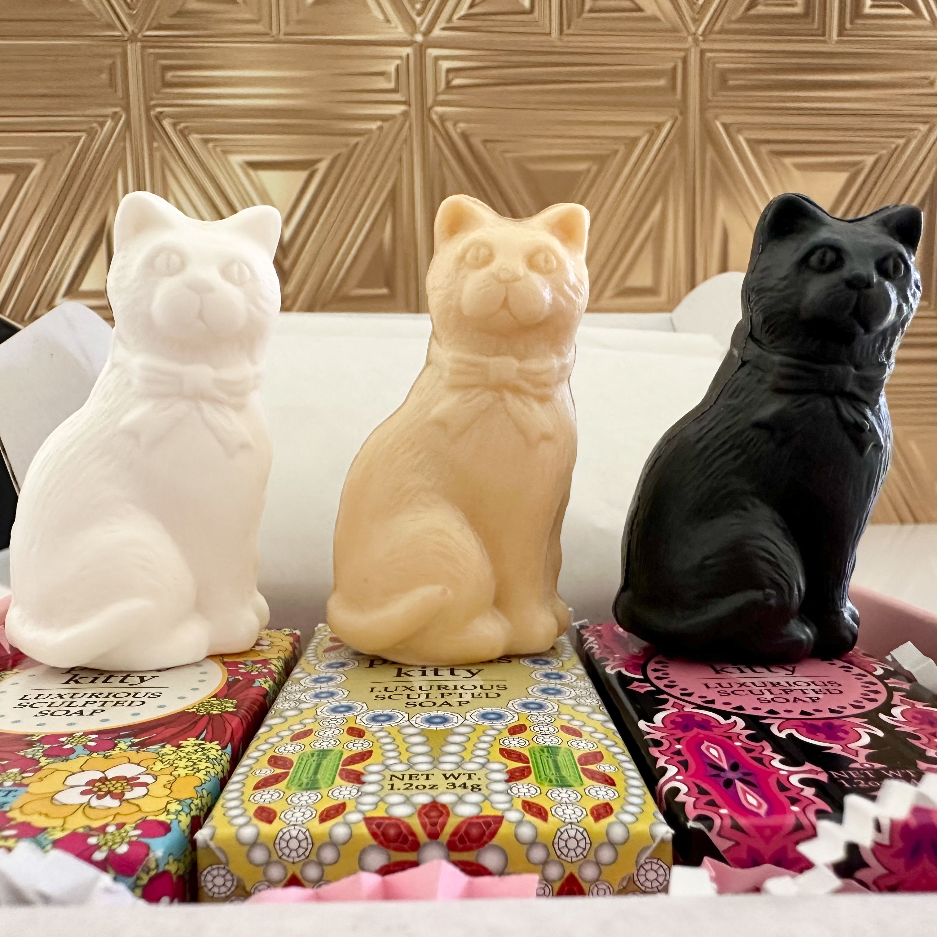 Three cat-shaped soap bars in white, beige, and black, displayed on top of their colorful patterned packaging in a gift box.