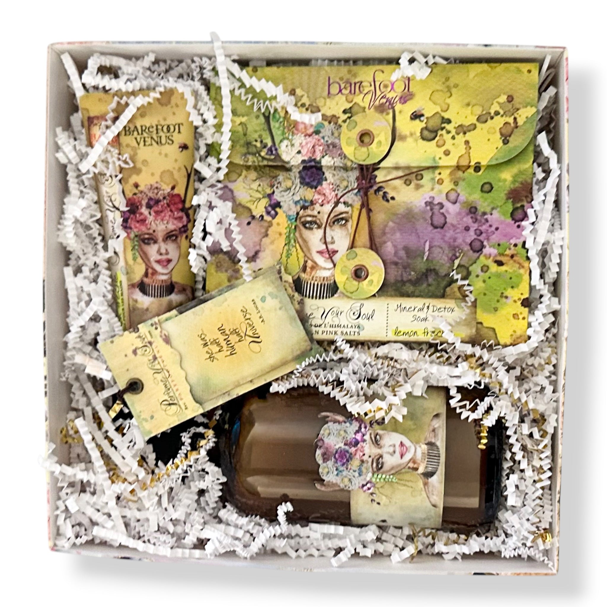 A curated gift box set containing a brown glass bottle, a tube of hair product, a bar of soap, and a decorative journal, all arranged in white shredded paper inside a square box. The items feature a consistent vintage, gothic aesthetic with illustrations of a woman's face and gothic text.