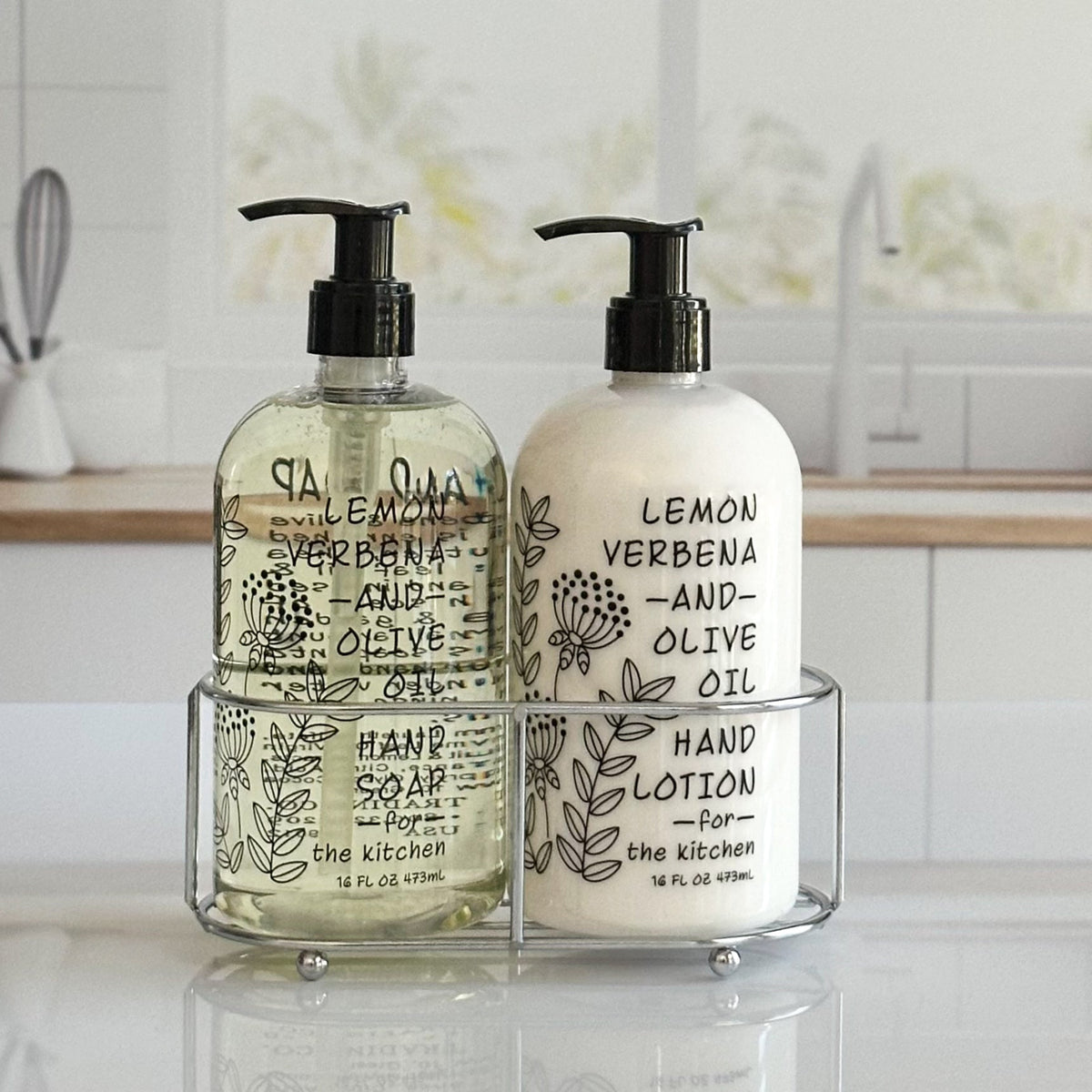 Two pump bottles of hand and body products displayed in a clear acrylic stand on a countertop. The left bottle contains pale yellow hand soap, and the right bottle contains white hand lotion, both with black pump tops and black botanical text labels reading lemon verbena and olive oil.