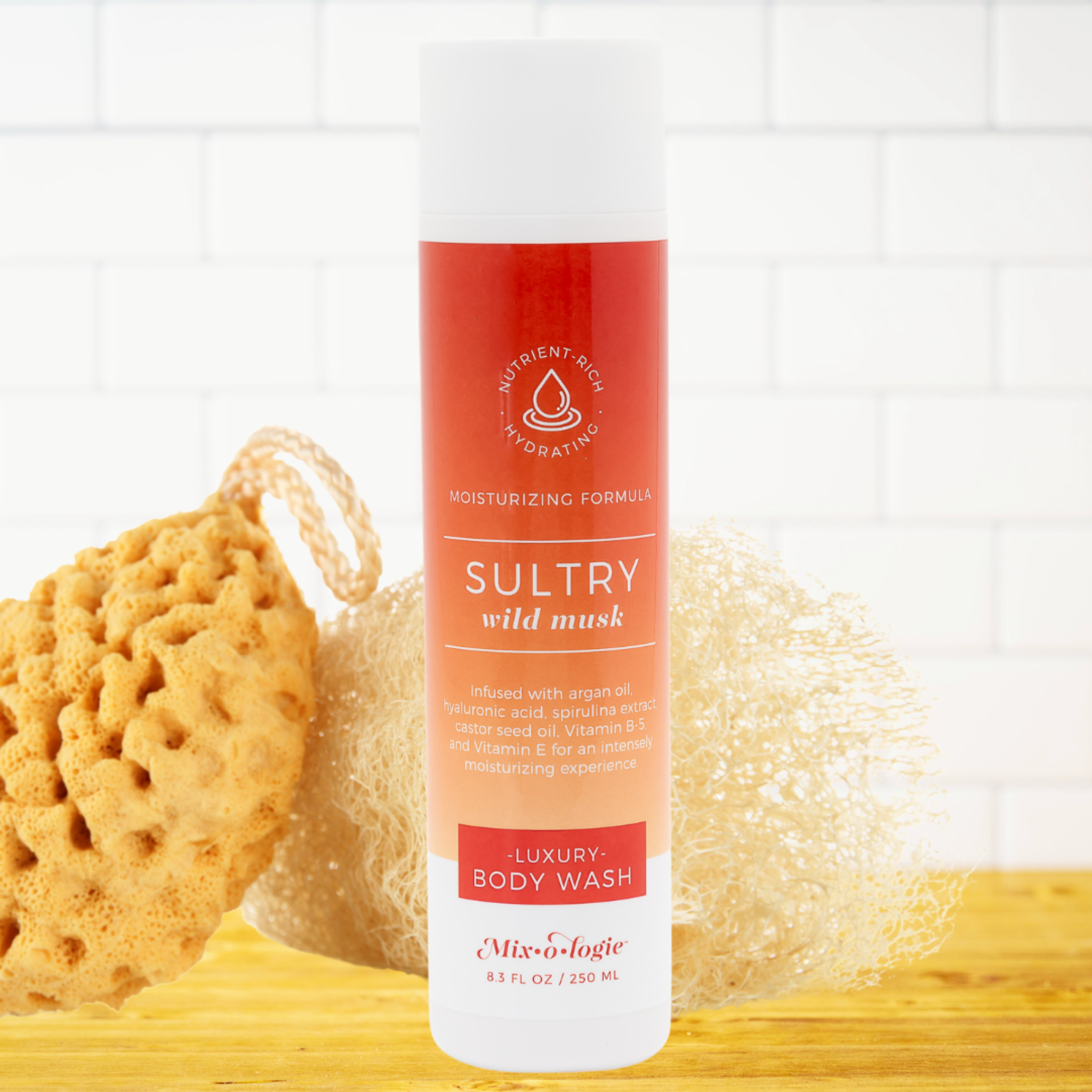 A bottle of Mix-It-logic Sultry Wild Musk Luxury Body Wash stands on a wooden surface with a natural loofah and a white mesh sponge behind it against a white subway tile wall.