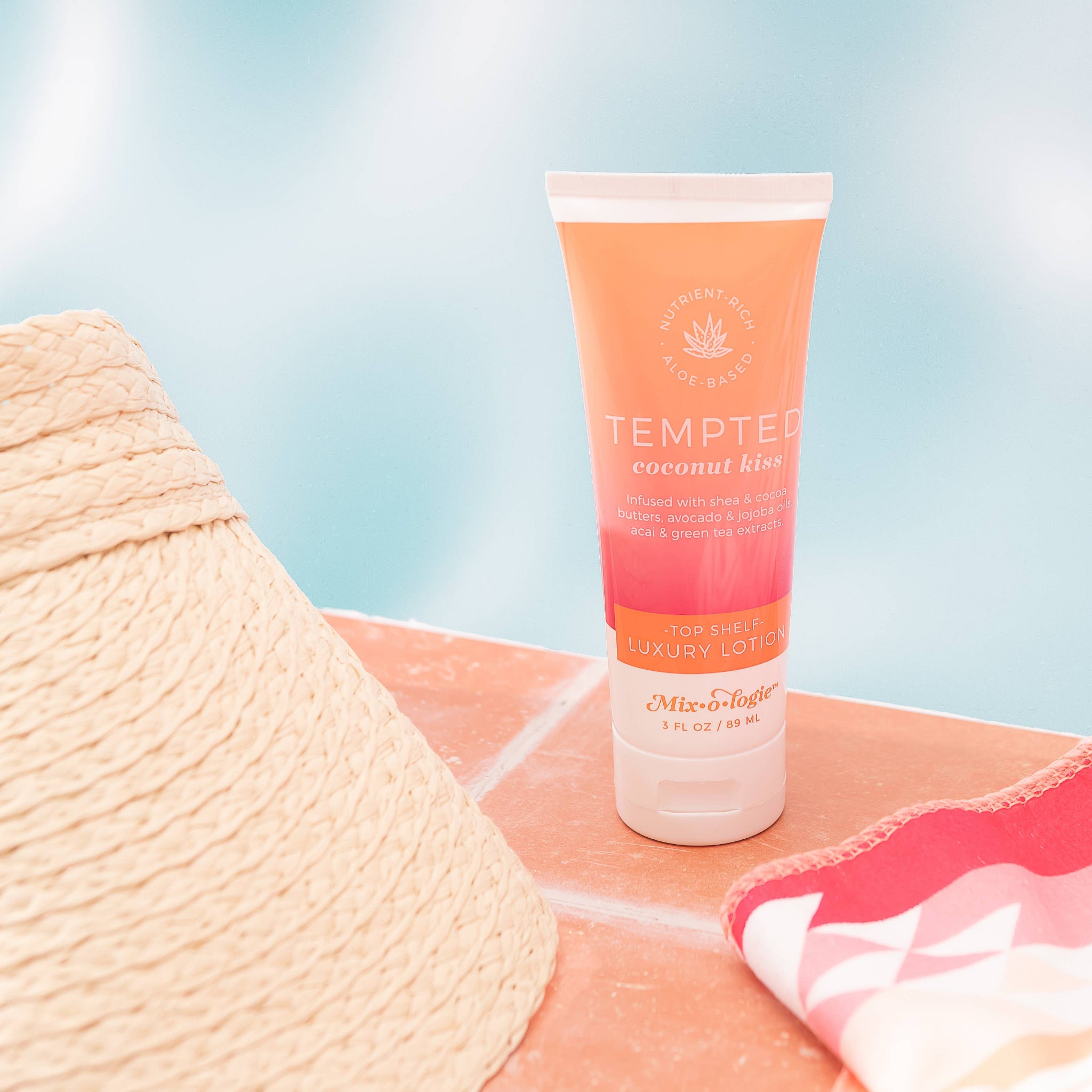 Tube of Tempted coconut glow lotion on a pink surface with a straw hat and towel in the background.