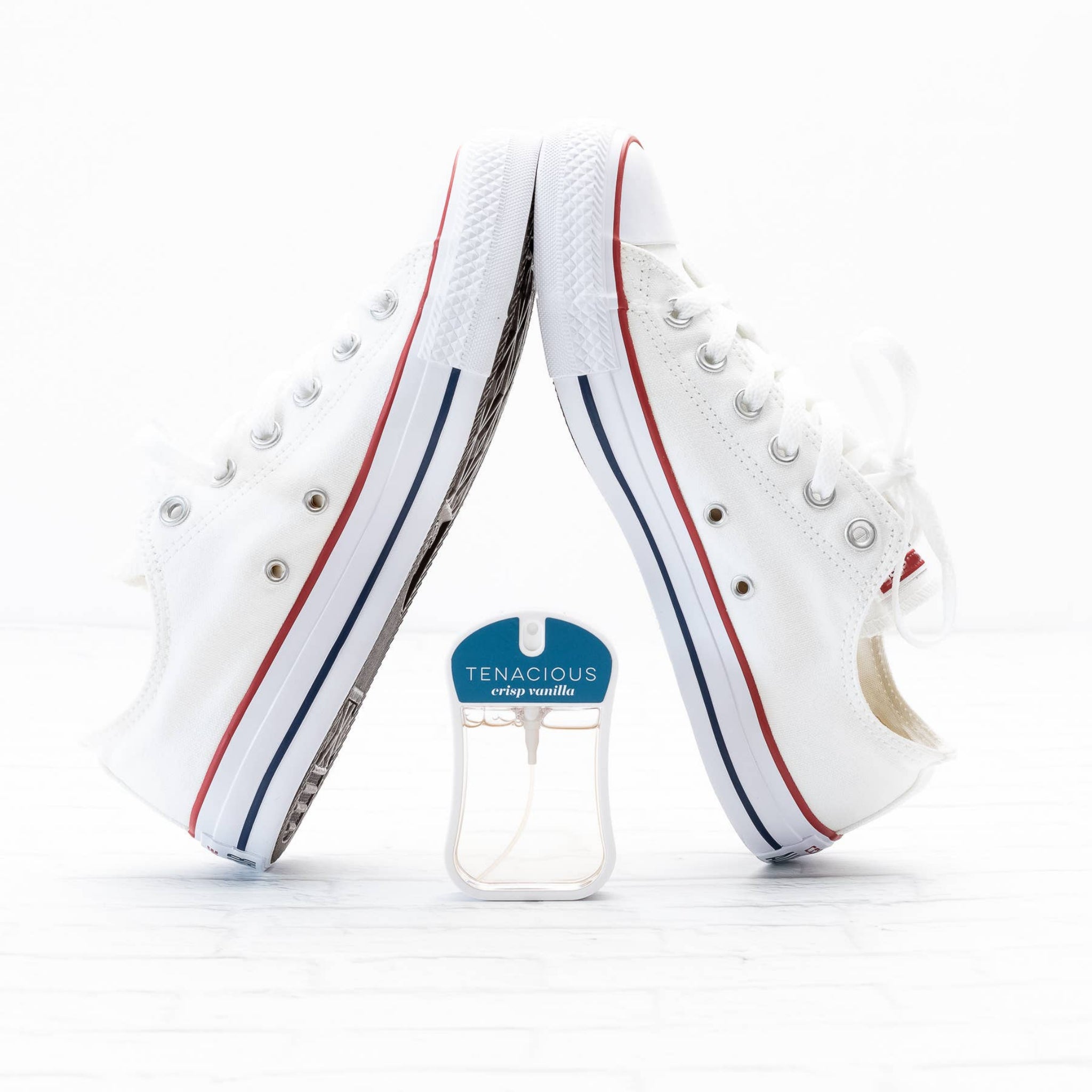 White sneakers with red and blue accents next to a Tenacious crisp vanilla air freshener on a white background