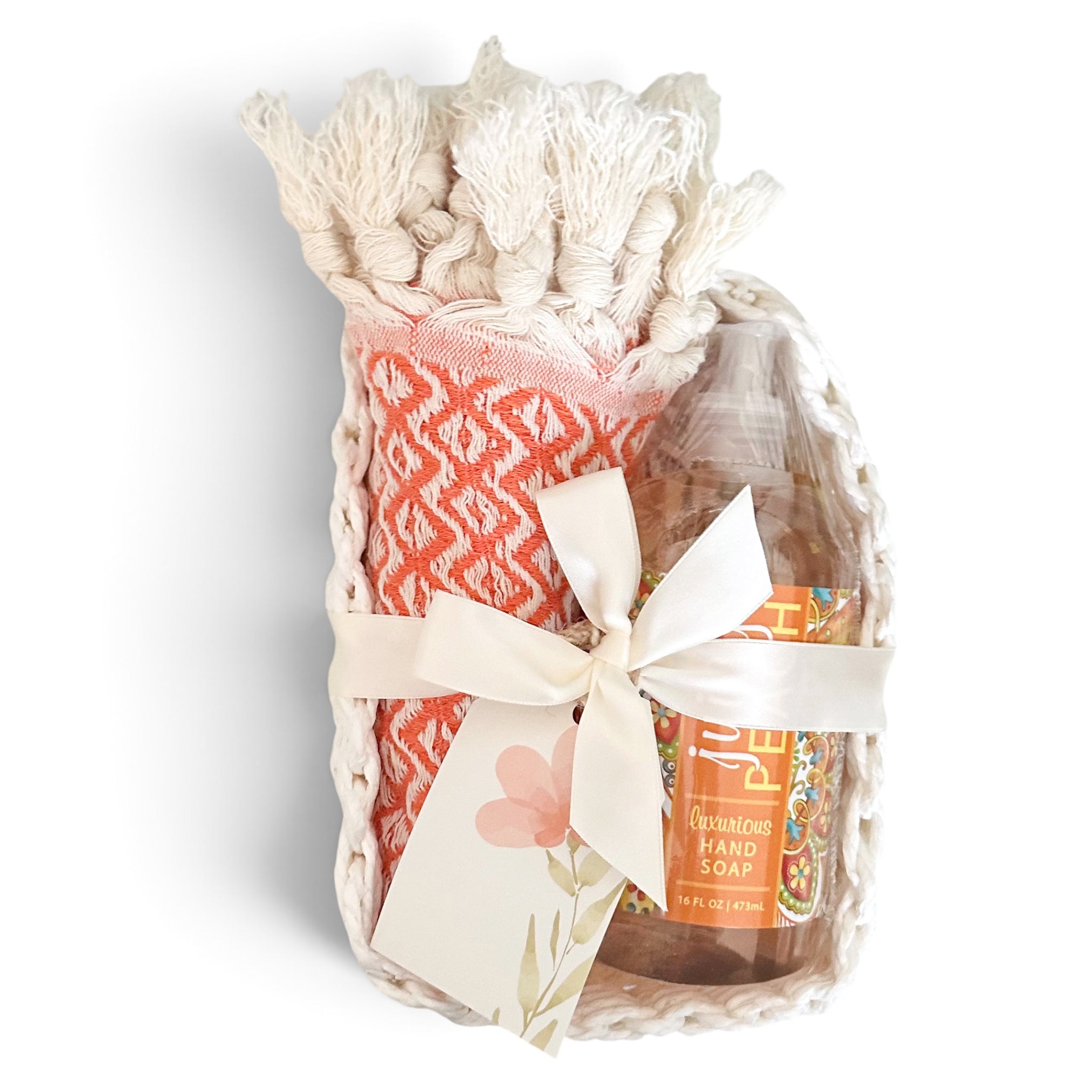 A gift set in a white woven basket containing a patterned cream and coral washcloth, a bottle of citrus hand soap, and a cream ribbon bow with a floral tag, on a white background.