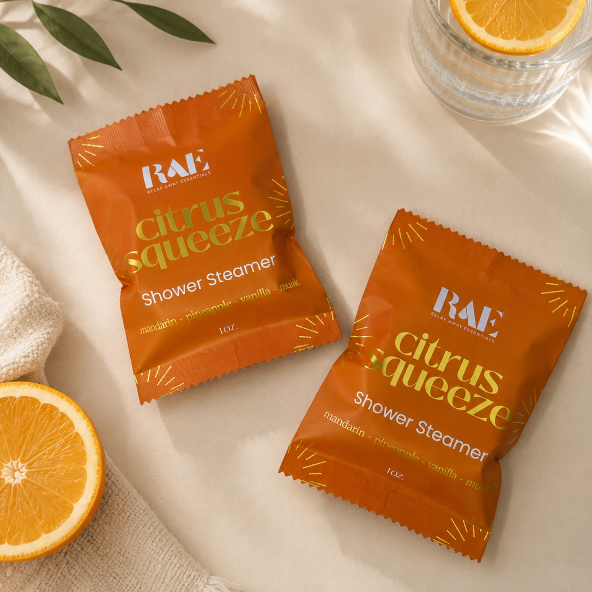 Two orange RAE Shower Steamer packets labeled 'citrus squeeze' arranged on a light beige surface with soft shadows, alongside a glass of water with an orange slice, a folded cream towel, and green leaves.
