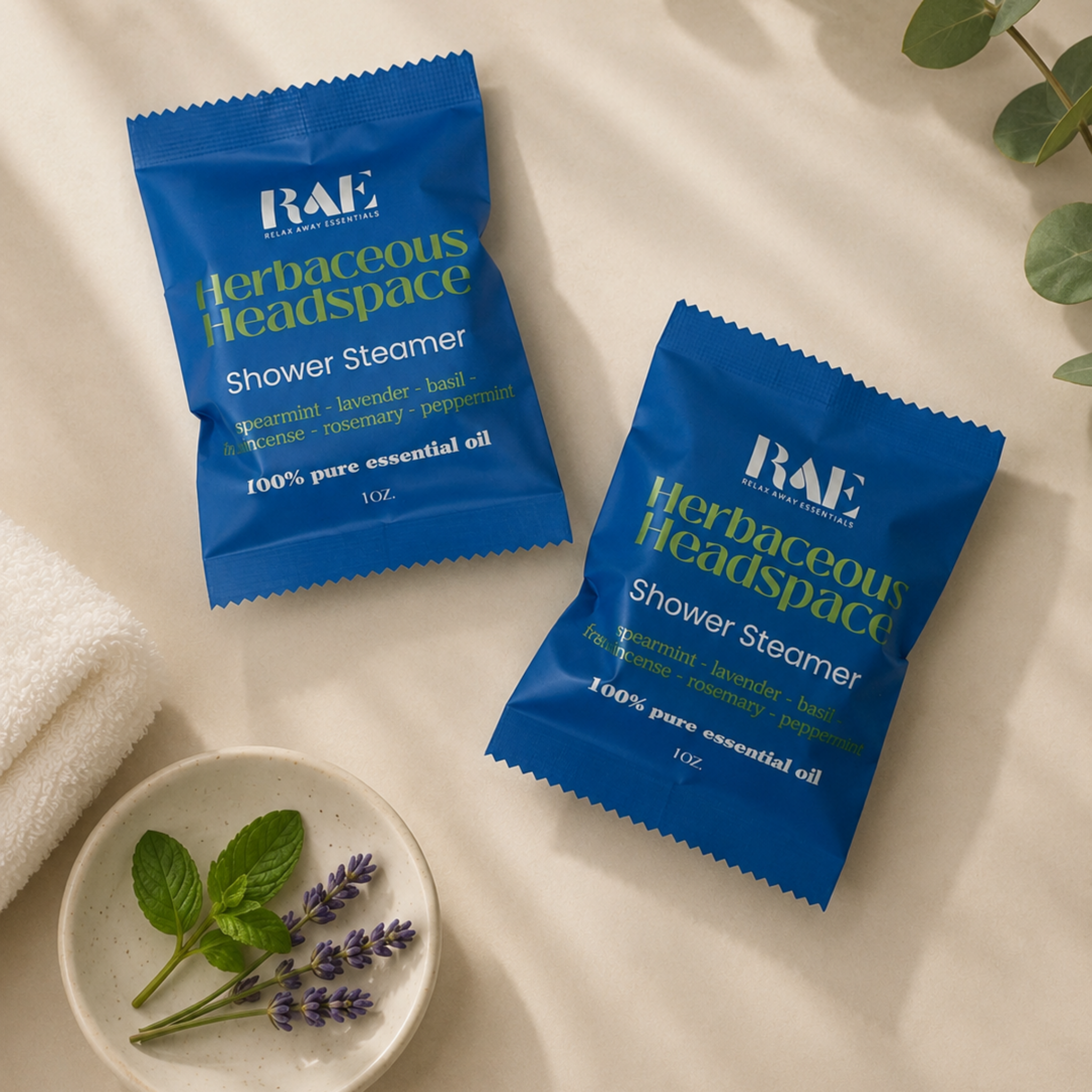 Two blue RAE Herbaceous Headspace shower steamer packets on a light surface with a small dish of lavender and mint leaves.