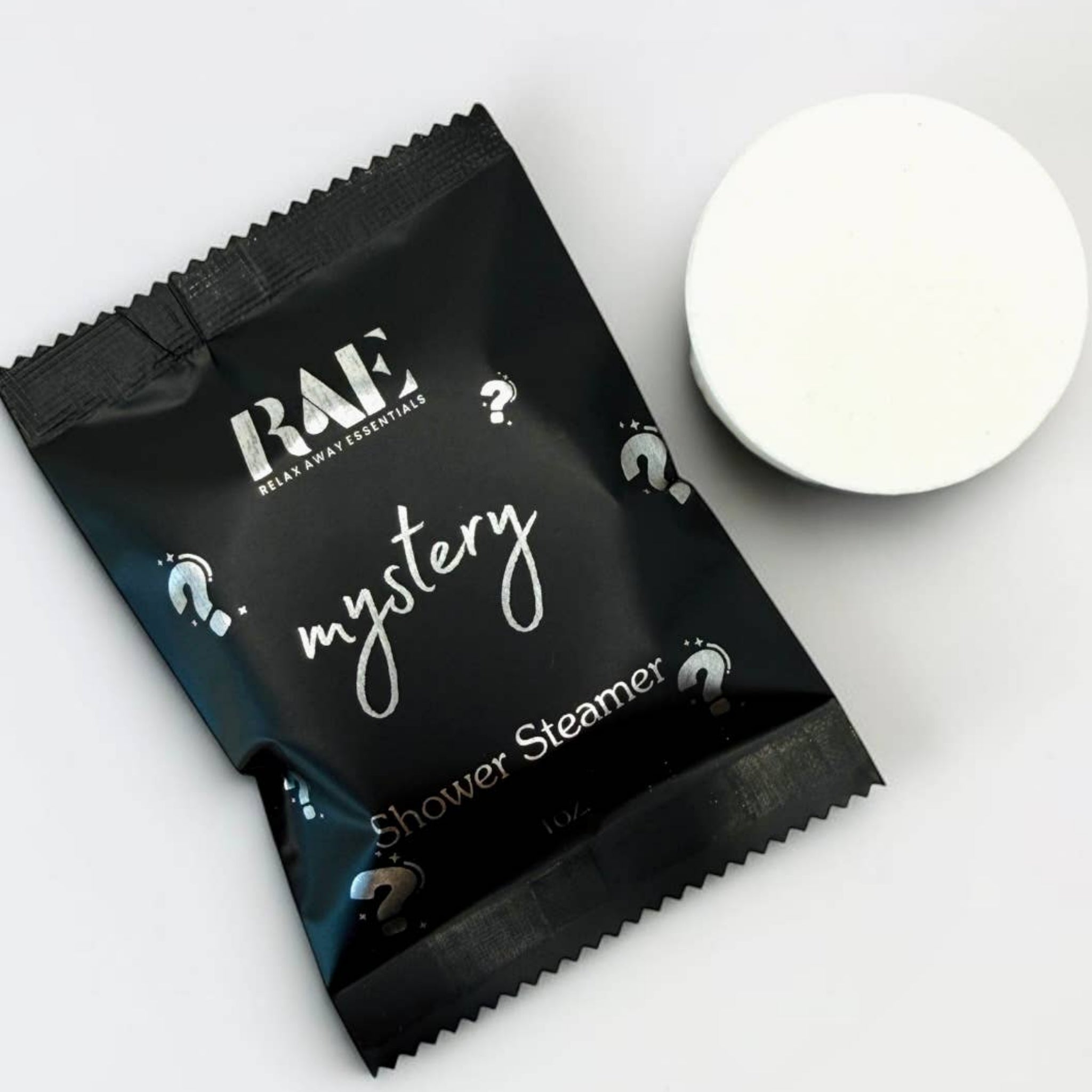 RAE mystery shower steamer packet with white steamer disc; black sachet on its side