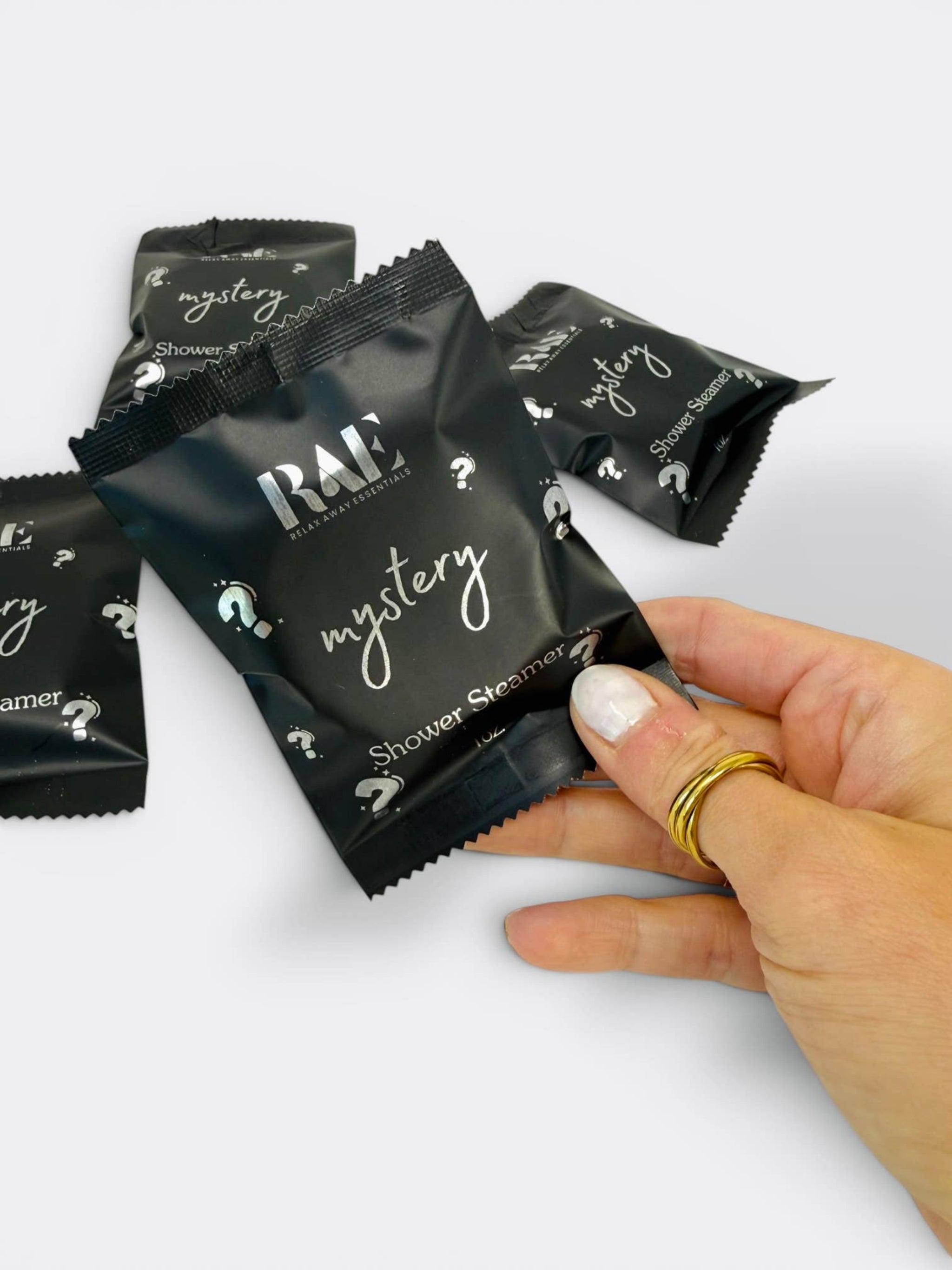 Hand holding RAE mystery shower steamer packet, black sachet with mystery name and 1 oz