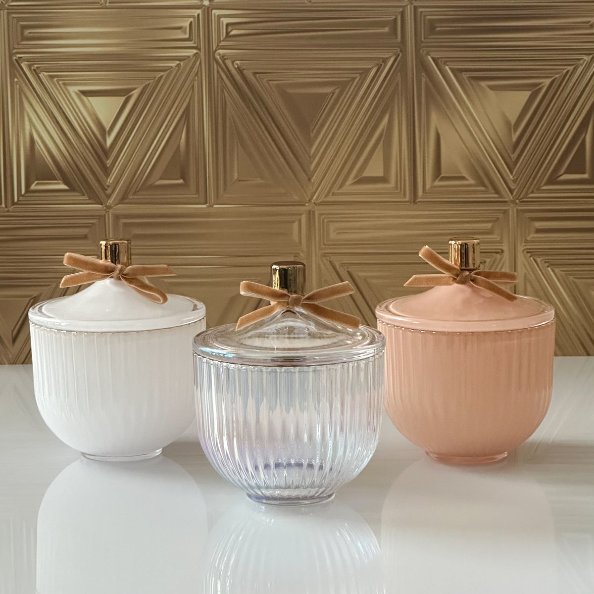 Three ribbed lidded jars with gold-tone knobs and bow details, shown in white, clear, and blush pink on a glossy white surface against a textured gold wall.