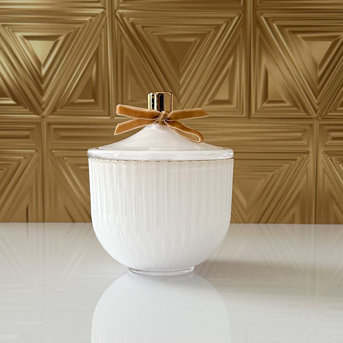 A white ribbed ceramic jar with a matching lid, decorated with a gold ribbon bow and a gold accent ring, set against a textured gold geometric wall.