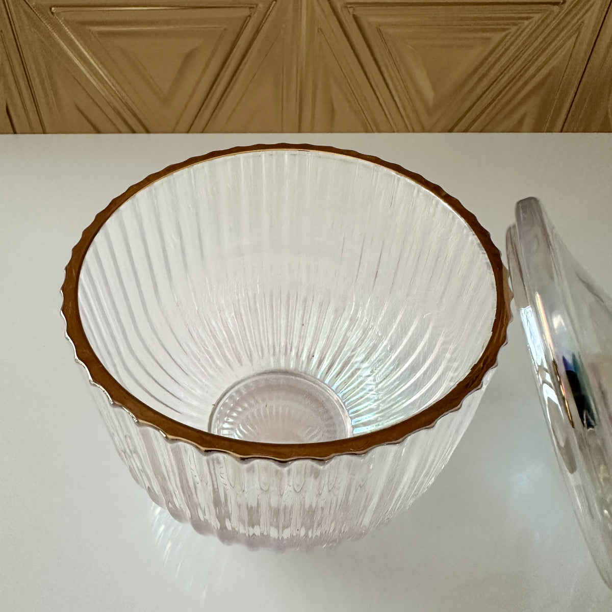 A clear ribbed glass bowl with a scalloped rim and a metallic gold edge, shown on a white surface with a matching clear glass lid partially visible on the right.
