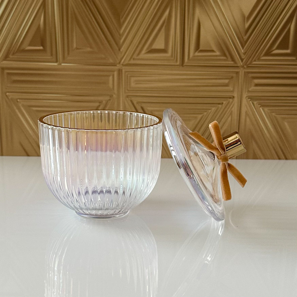 A clear ribbed glass bowl with a matching glass lid, accented with a gold rim and a small gold bow, set on a white reflective surface against a textured gold wall.